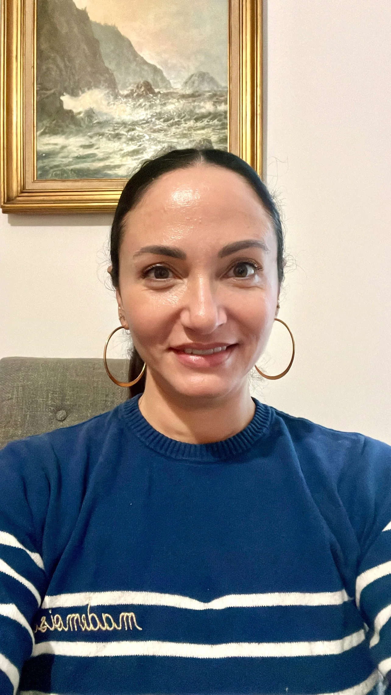 A woman taking a selfie indoors, wearing a blue and white striped sweater with cursive writing, gold hoop earrings, and has dark hair slicked back. A painting of a seascape with waves crashing on rocks is visible in the background.