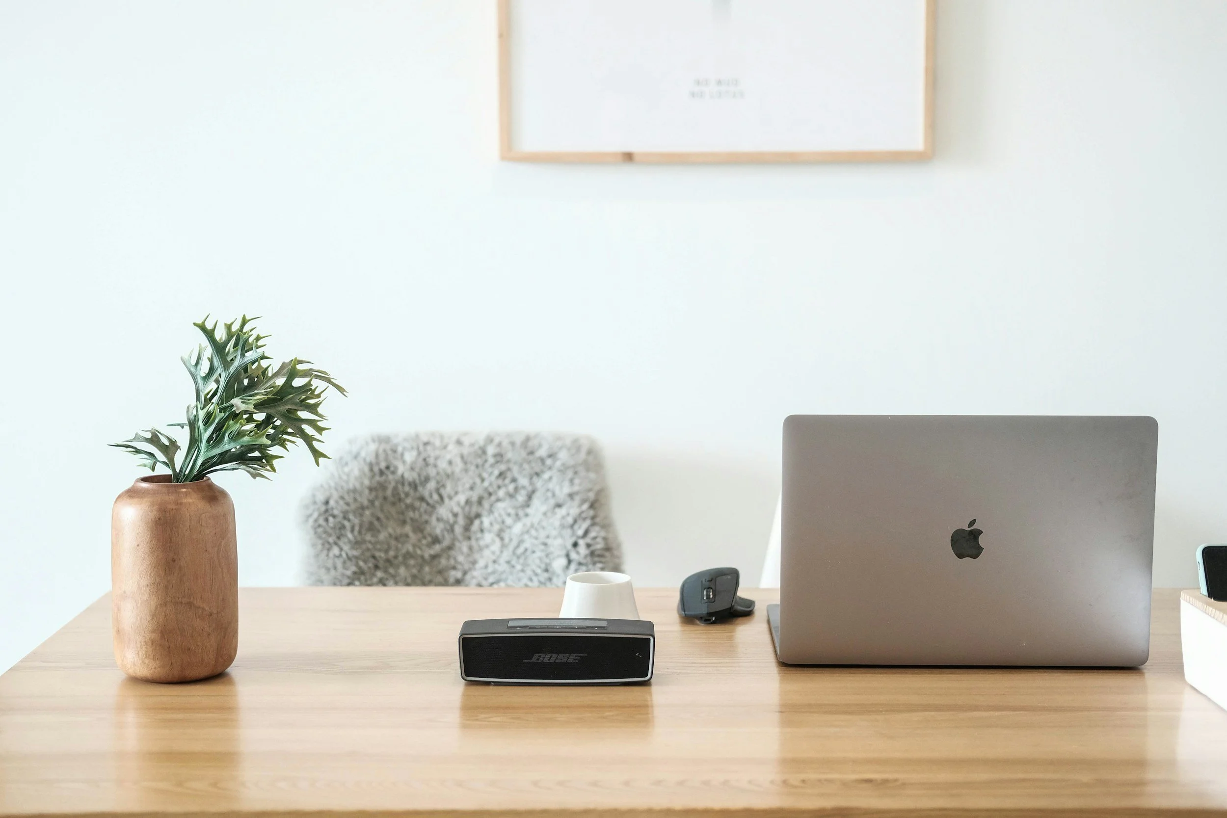 A wooden desk with a potted plant in a terracotta vase, a Bose speaker, a white mug, a black cell phone, a closed laptop, and a small notepad. In the background, there is a white wall with a framed picture and a patterned chair.