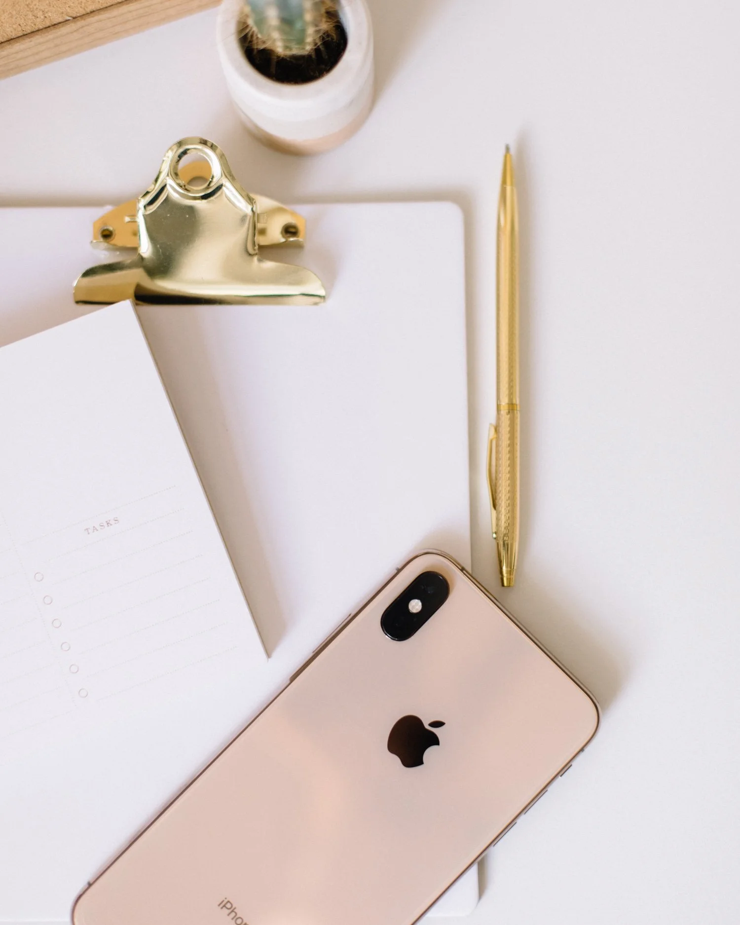 A gold iPhone with black Apple logo, a gold pen, a clipboard with blank paper, a cactus in a white pot, and a pink iPhone with dual cameras on a white surface.