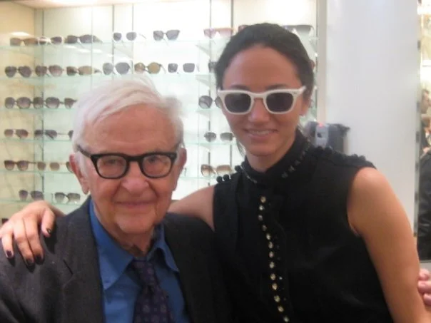 A young woman and an older man pose together in an eyewear store, wearing sunglasses. The woman is wearing large white sunglasses and a black sleeveless top with pearls, while the man is wearing black glasses, a suit, and a tie. Shelves of sunglasses are visible in the background.