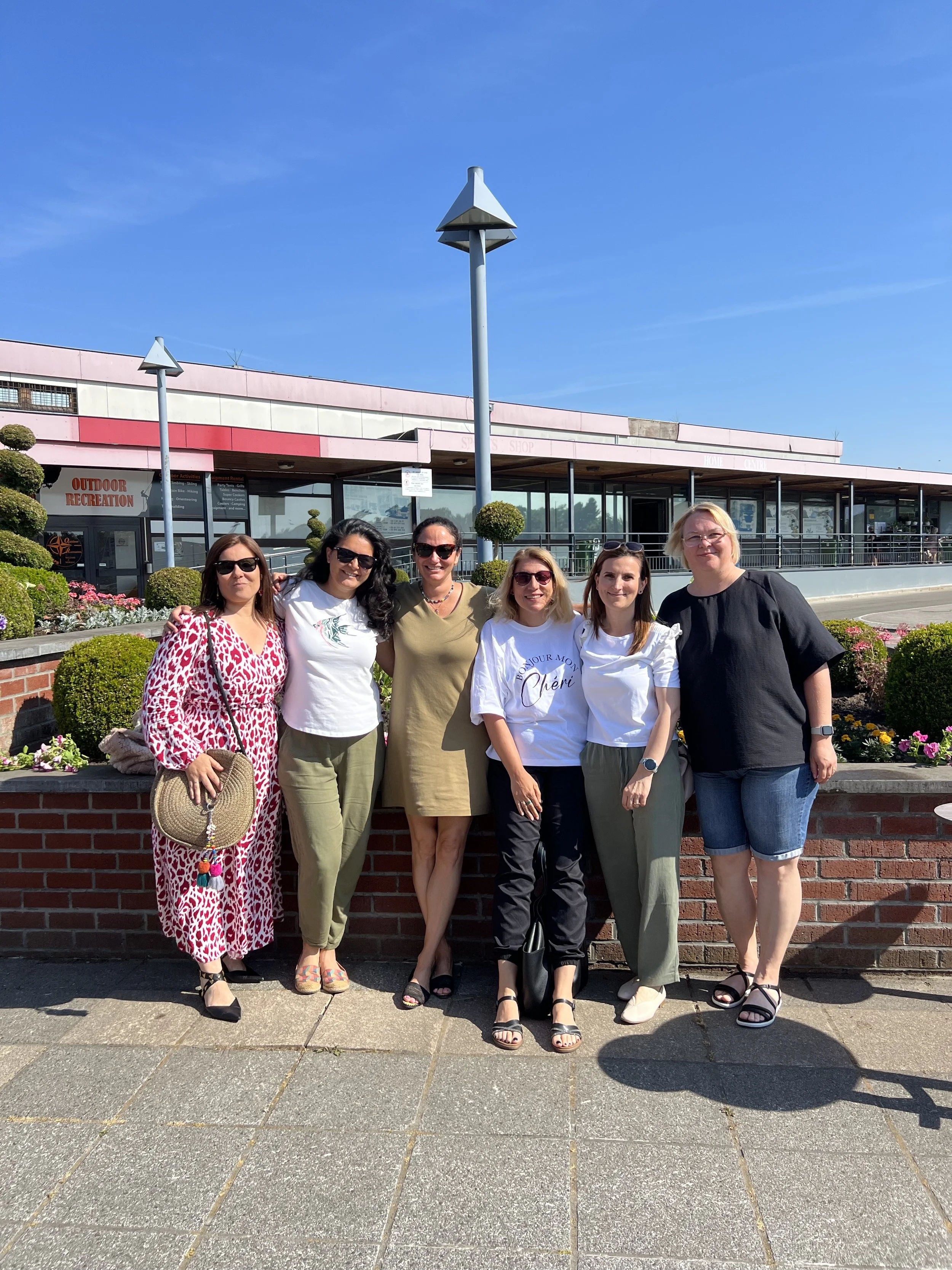 Six women standing in front of a building with a sign that reads 'OUTDOOR RECREATION.' They are outdoors on a sunny day, smiling and posing for the photo.