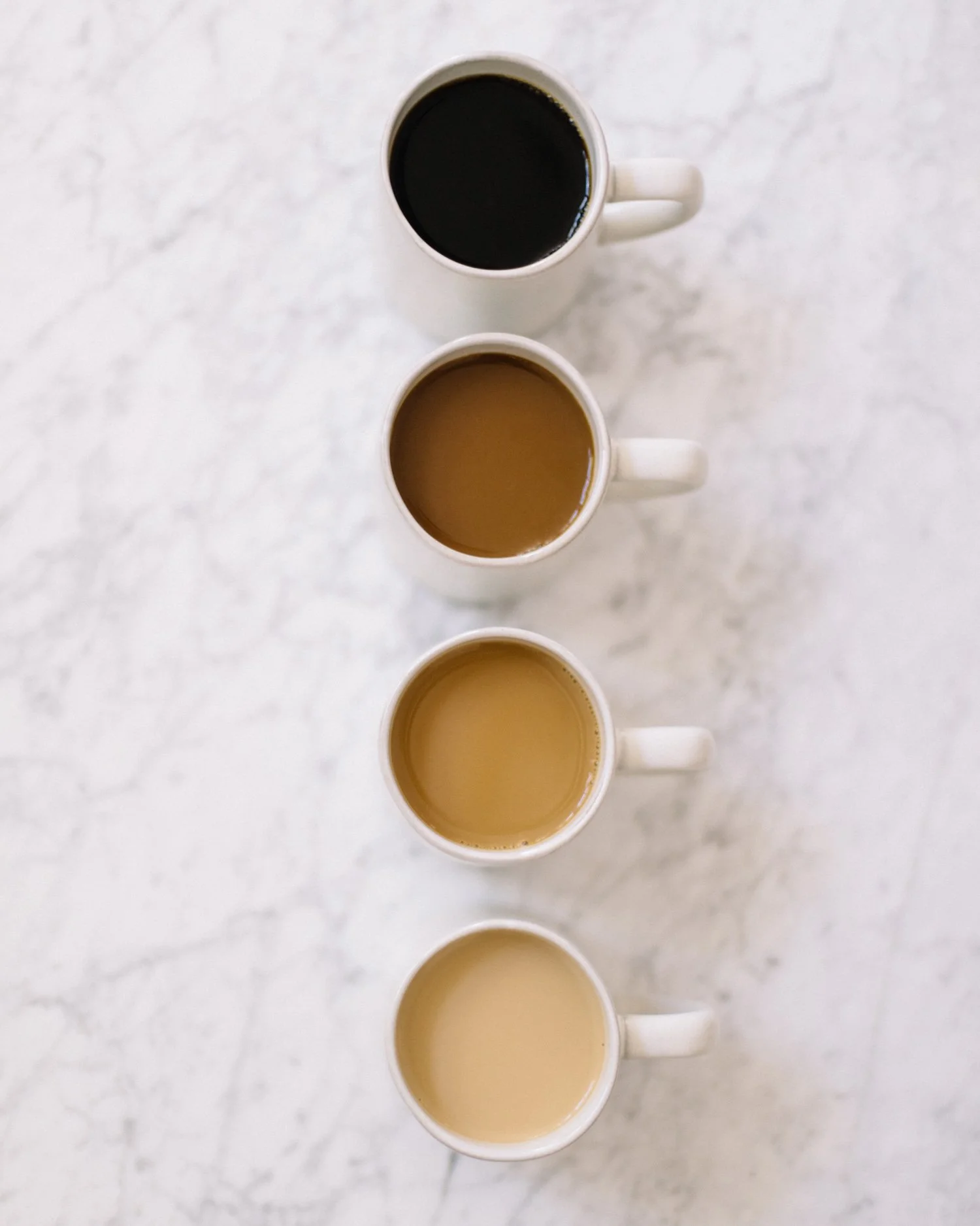 A top-down view of four white coffee mugs filled with different types of coffee or tea, arranged in a vertical line on a light marble surface.