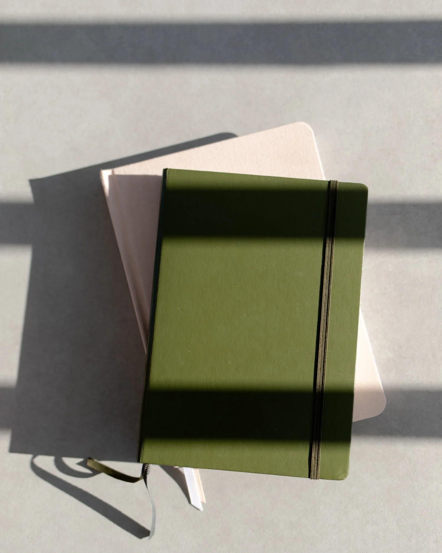 A green closed notebook with a black elastic band on a gray table, casting shadows from sunlight.