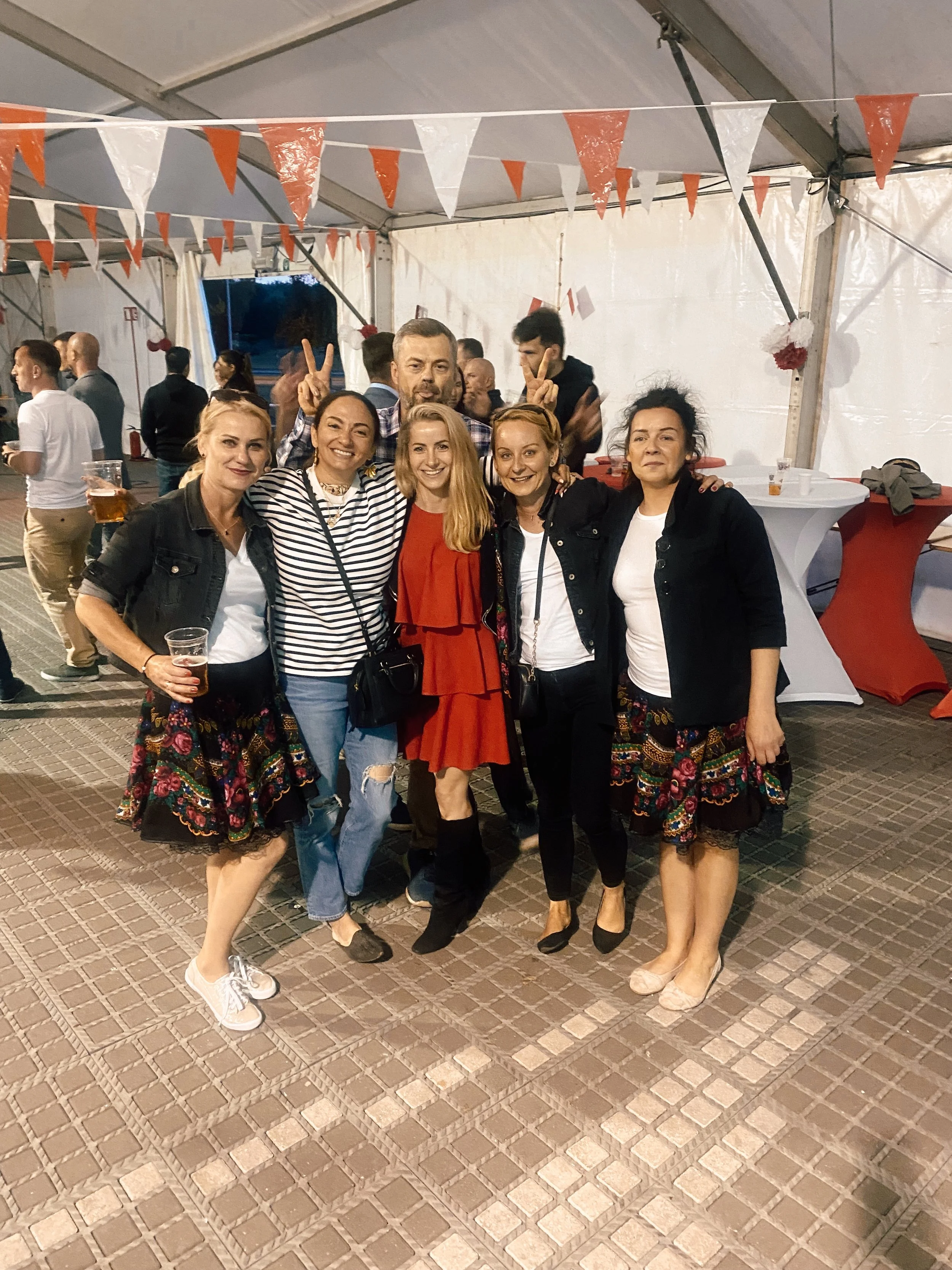 Group of five women and a man posing together at an indoor event, with some additional people in the background. The women are smiling and dressed casually, and one woman is holding a drink. The background has decorations hanging from the ceiling and