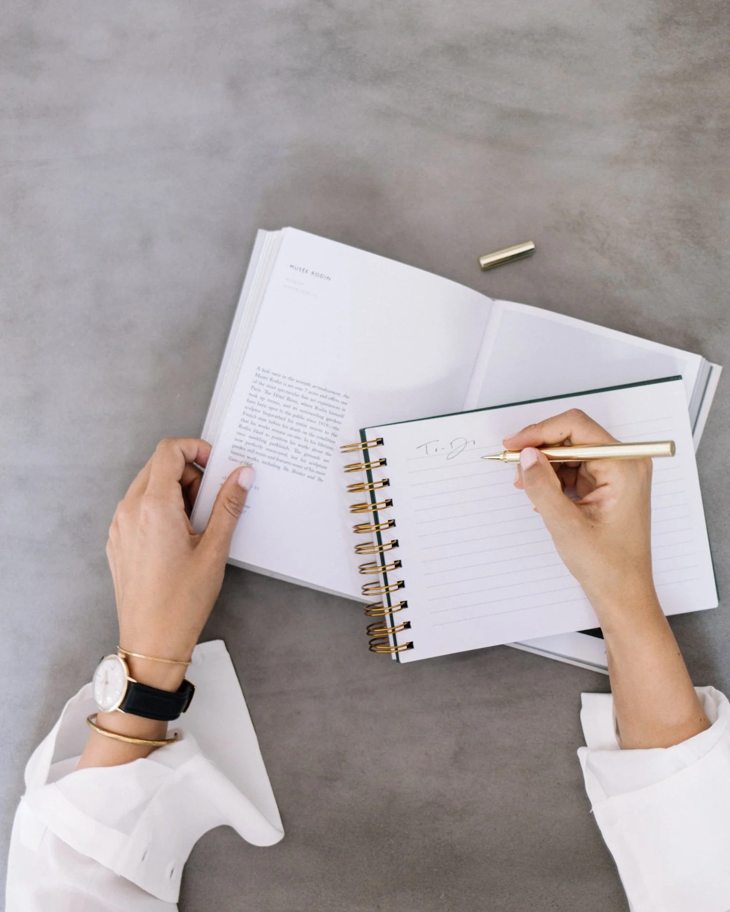 A person wearing a white long sleeve shirt, a watch, and bracelets is writing on a lined notepad with a gold pen. There is an open book or magazine on the table and a gold pen lying on the table.