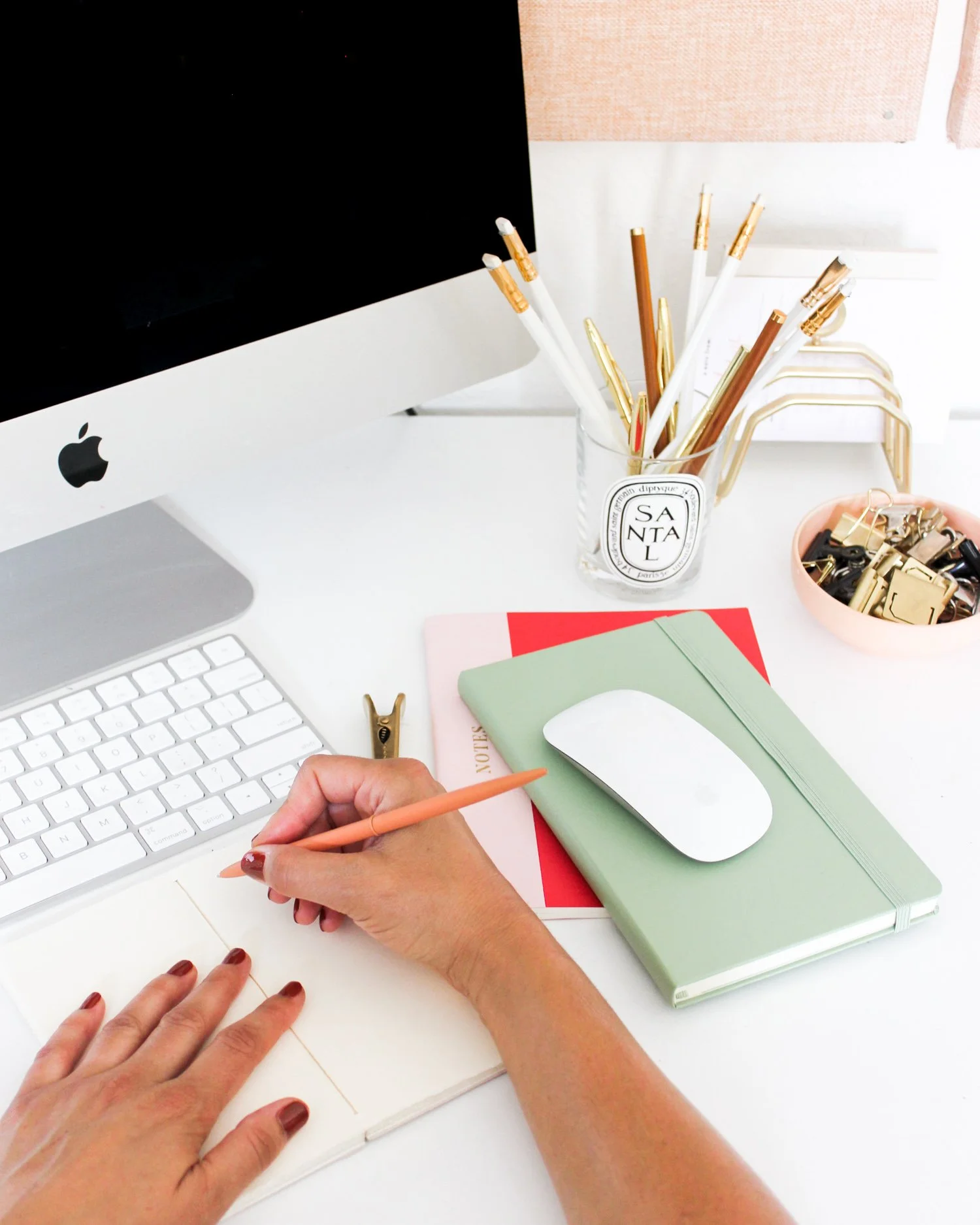 A person writing with a peach-colored pencil on an open notebook, with a white wireless mouse, and an iMac computer on a white desk. The desk also has notebooks, a glass container with pens, and a pink dish with gold paper clips.