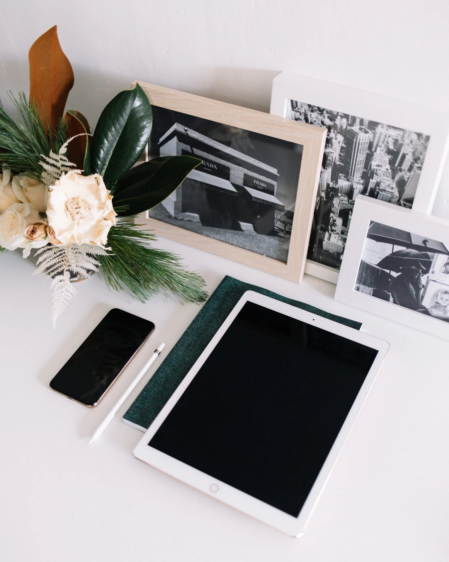 A white desk with a smartphone, a tablet, a stylus, framed black and white photos, and a bouquet of flowers and greenery.