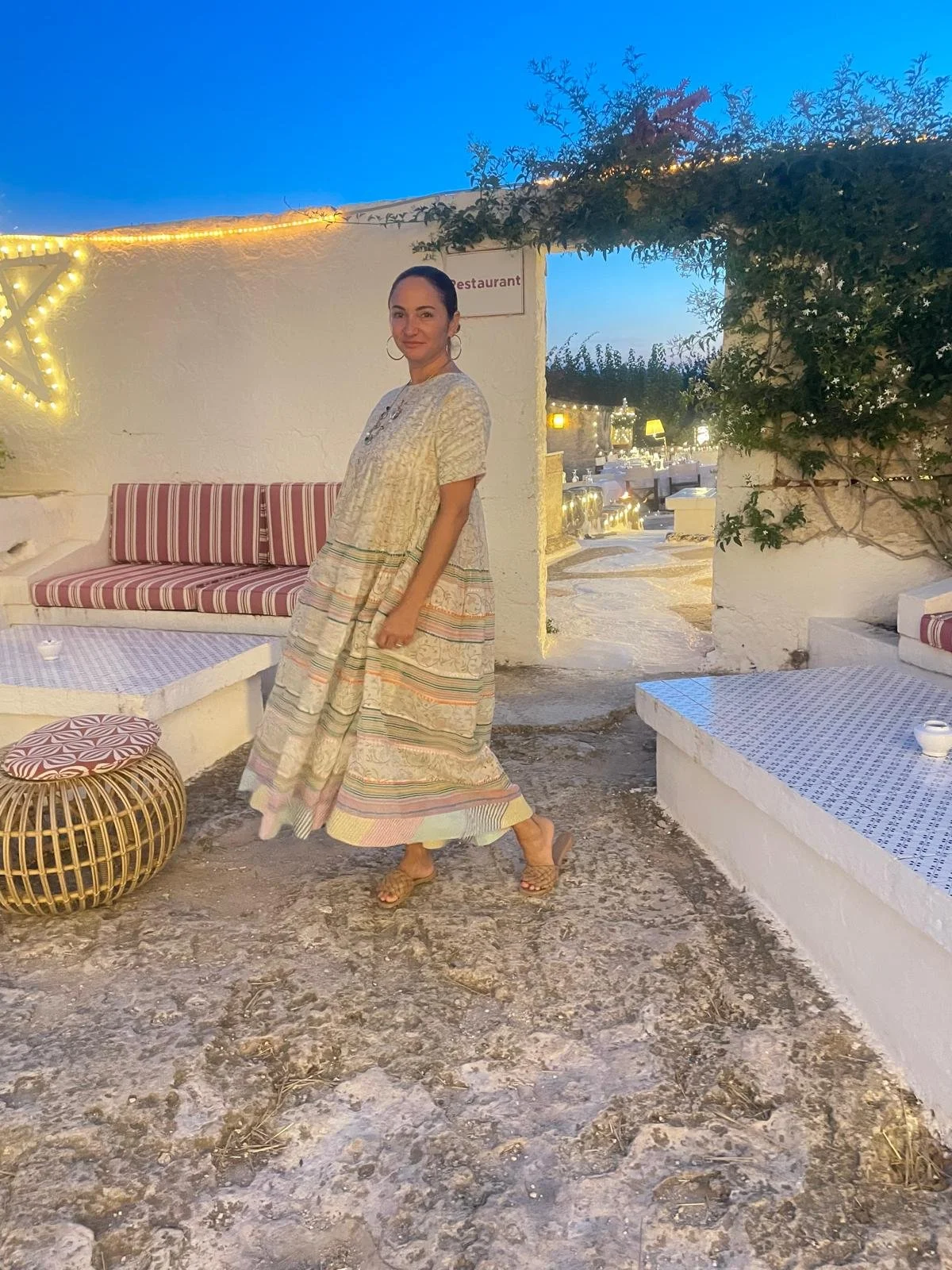 Woman in a flowing, patterned dress inside a Mediterranean-style outdoor restaurant or lounge area, with white walls, string lights, and outdoor furniture, during twilight.
