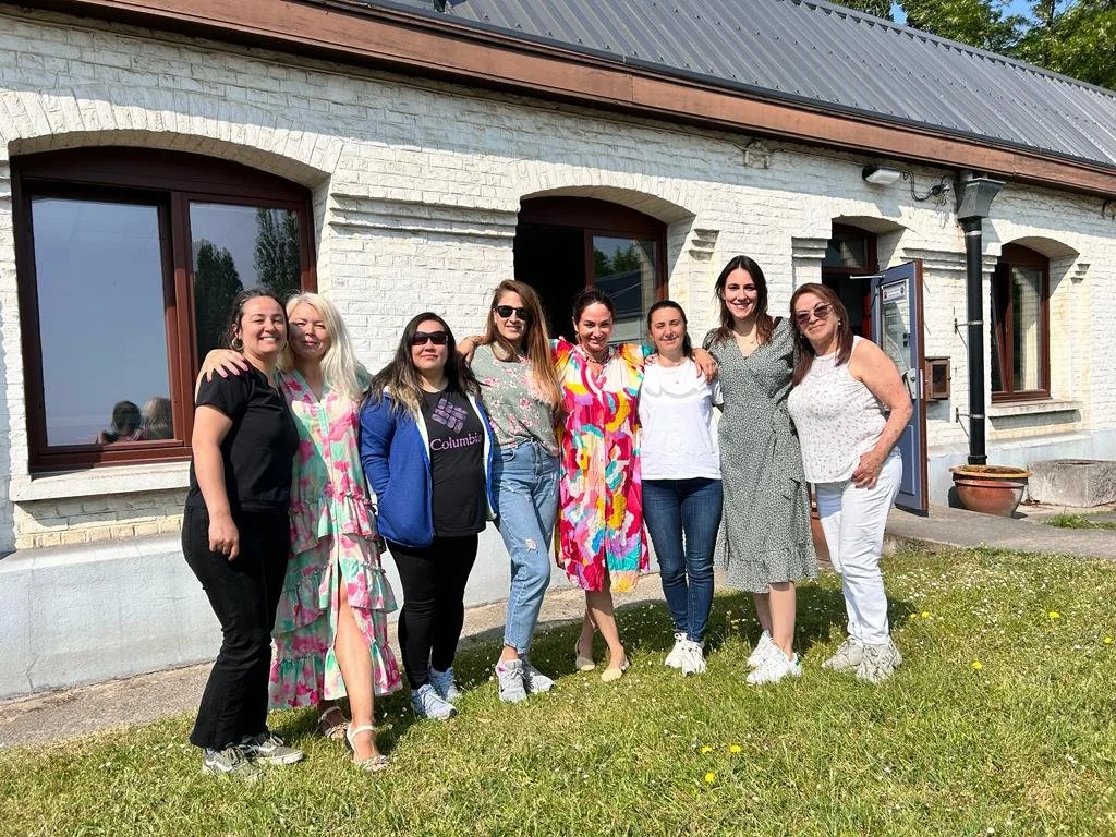 Group of eight women standing outside in front of a white brick house on a sunny day, smiling and posing together.