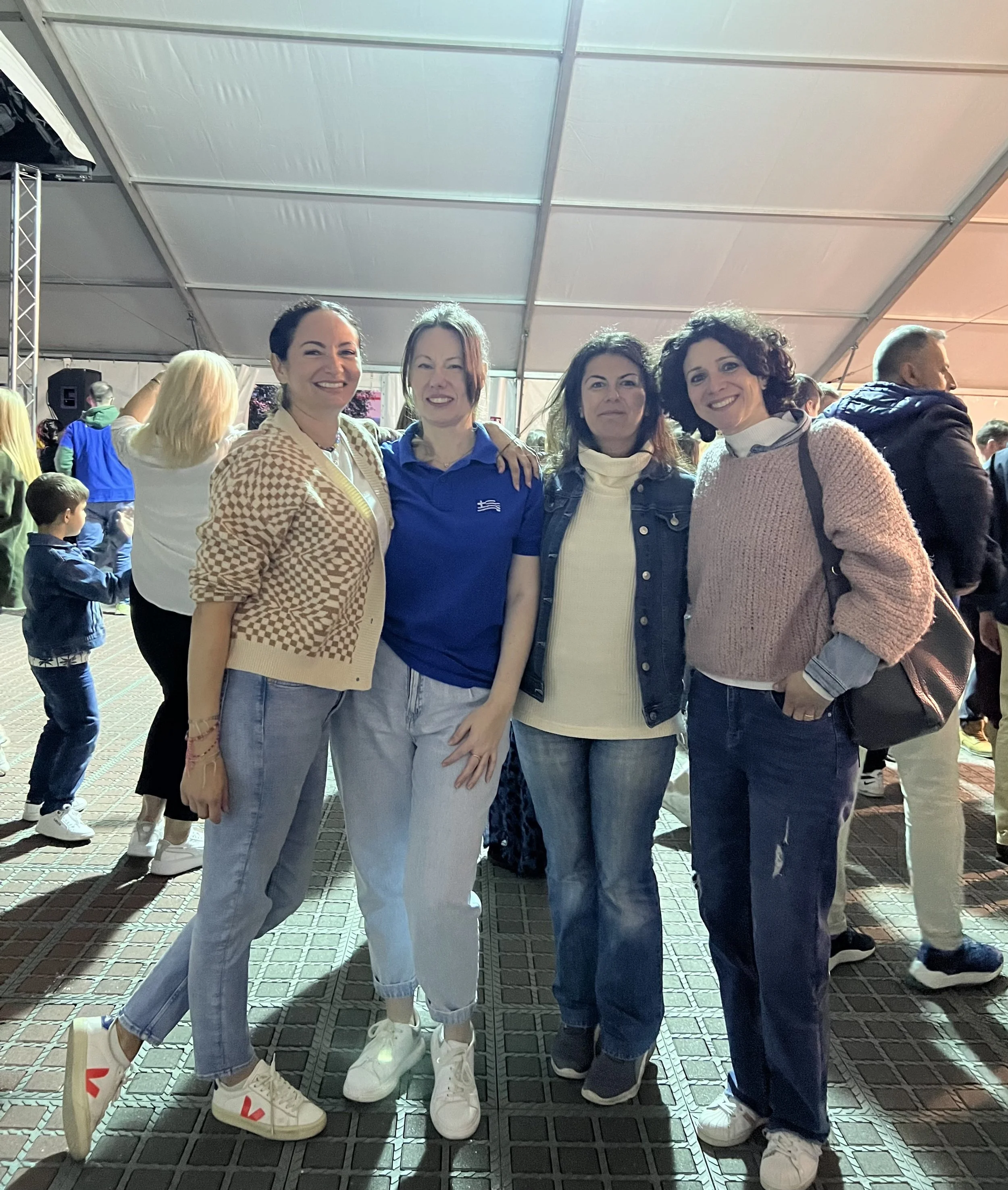 Four women standing together in an indoor event space, smiling at the camera. They are casually dressed, and there are other people in the background, with a large tent overhead and a tiled floor.