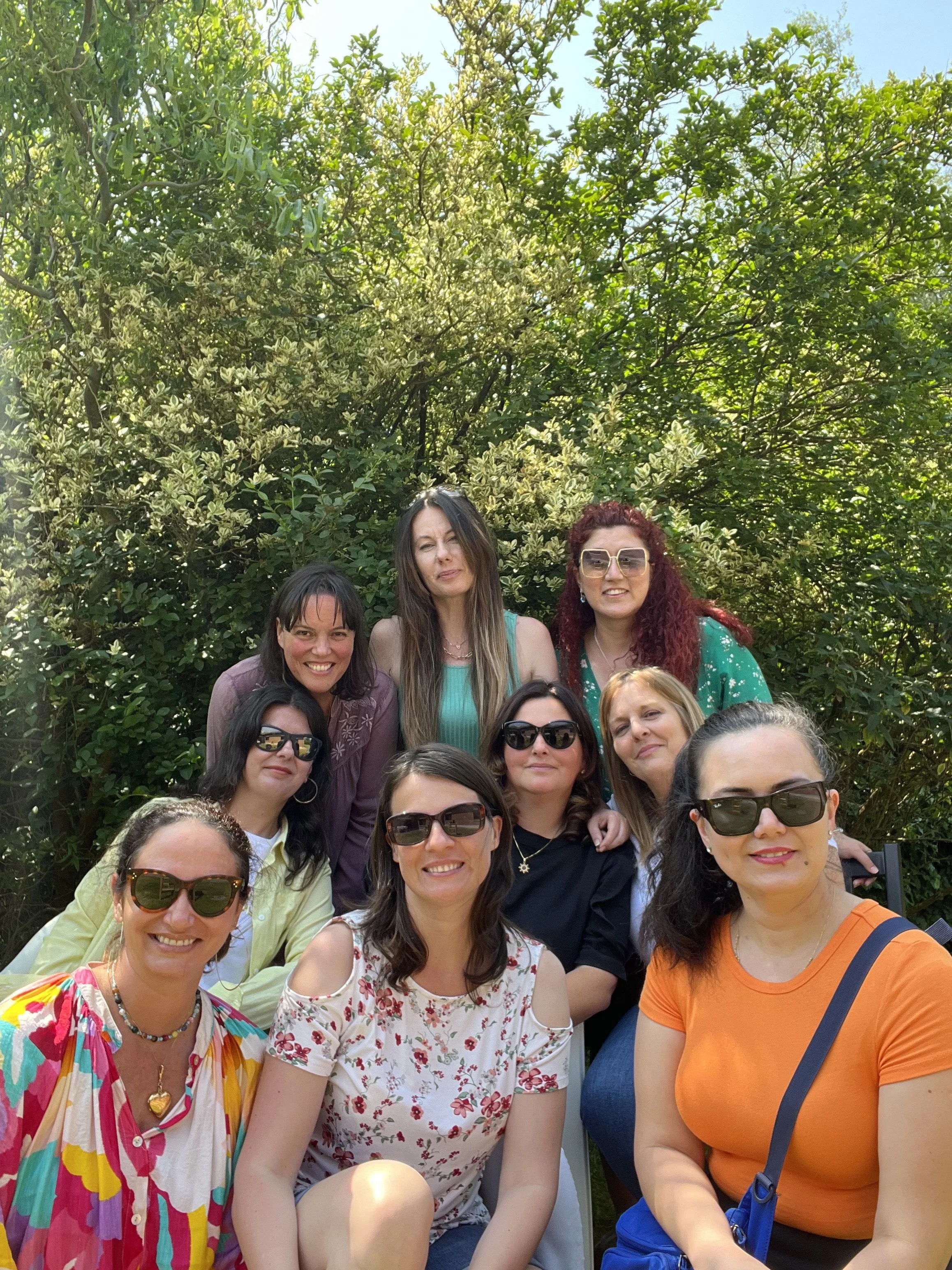 A group of women gathered outdoors for a photo, standing in front of lush green trees and bushes on a sunny day.