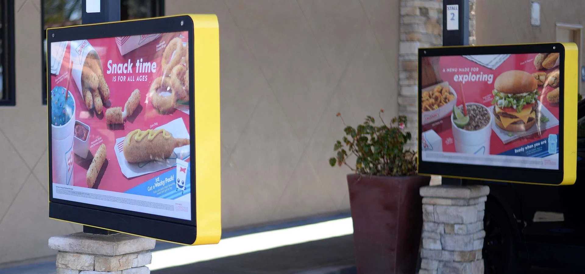 Digital menu screens outside a fast-food restaurant, with food images and promotional text for snacks, burgers, and drinks.