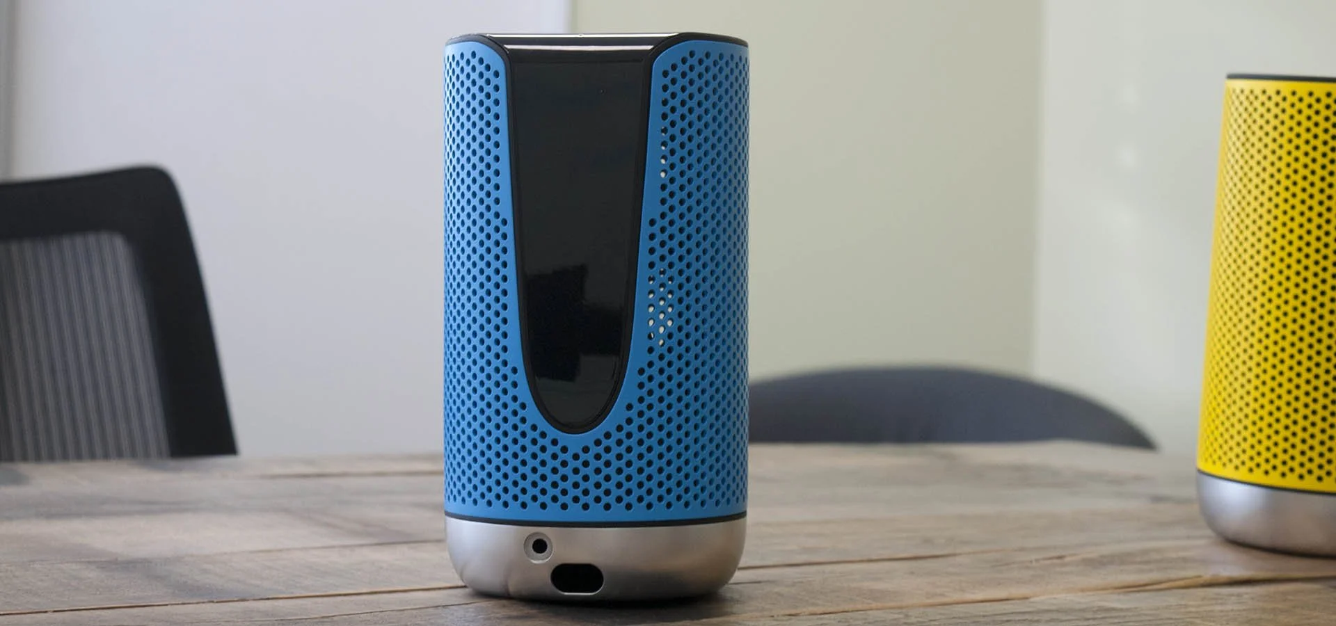 Blue and black cylindrical smart speaker on a wooden table, with a yellow similar speaker partially visible on the right.