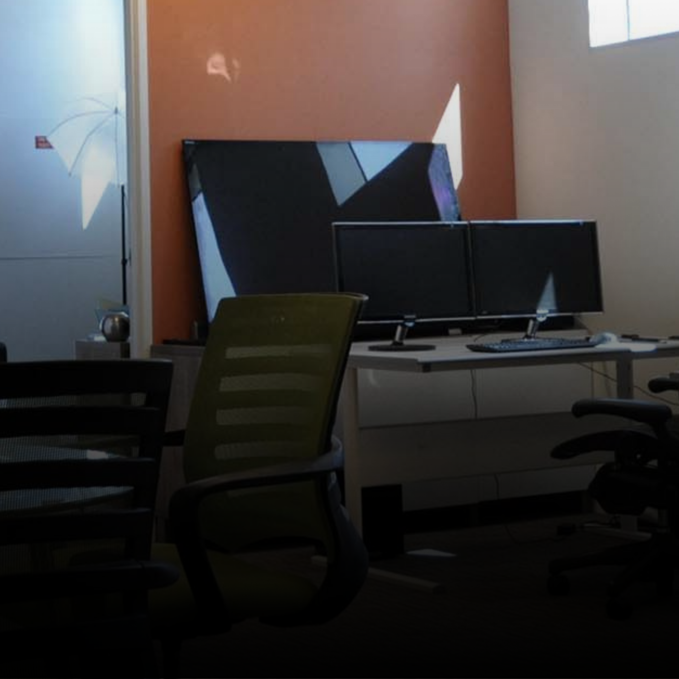 Office workspace with a desk holding two monitors, a keyboard, and a mouse, alongside office chairs, and a wall with artwork.