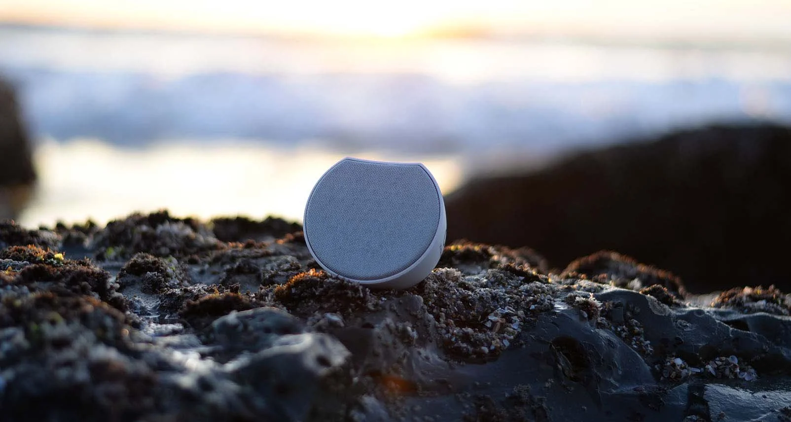 Wireless speaker on a rocky beach during sunset with ocean waves in the background.