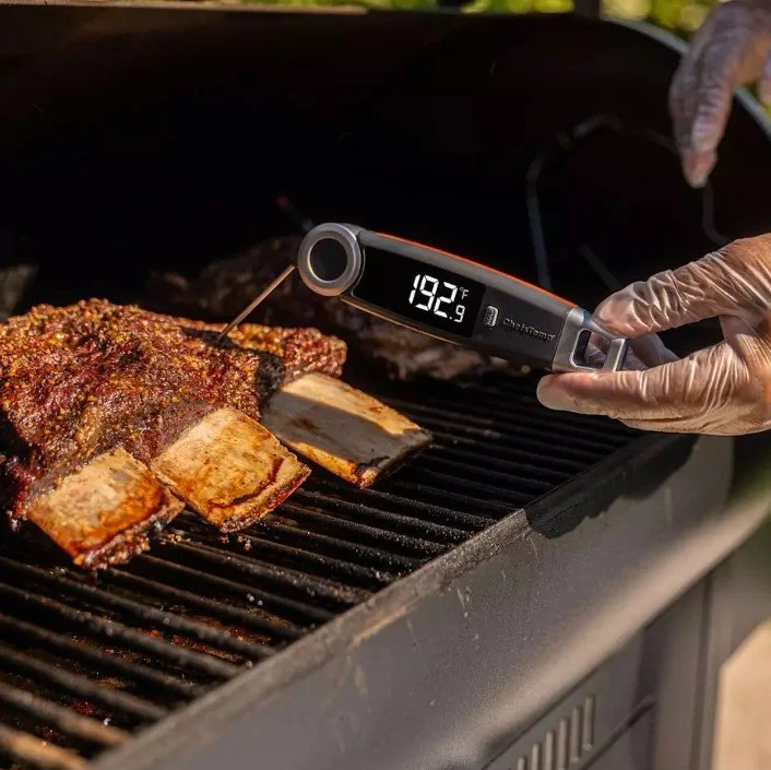 Close-up of a meat smoker grill with glazed pork ribs being checked for temperature, showing 192.9°F on a digital meat thermometer, with gloved hand holding the thermometer over the ribs.