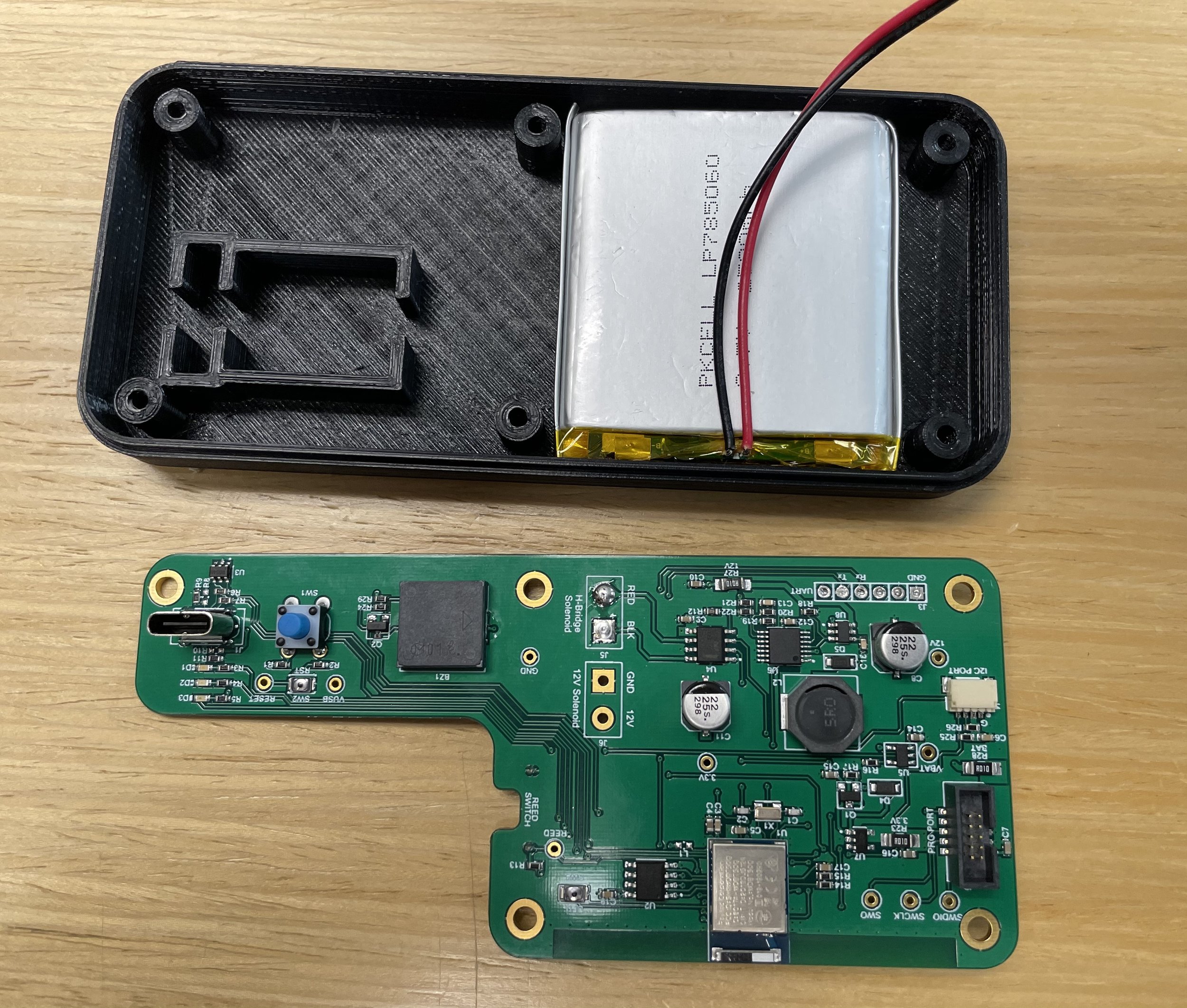 Open black plastic electronic enclosure with a green circuit board, a silver rectangular component, and wires inside, placed on a wooden surface.