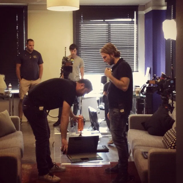 Group of four men in a room with cameras, lighting, and equipment. One man is bending over a laptop on the floor, another is standing near him, and two others are observing near the back. The room has window blinds, sofas, and a table with bottles and coffee in the background.