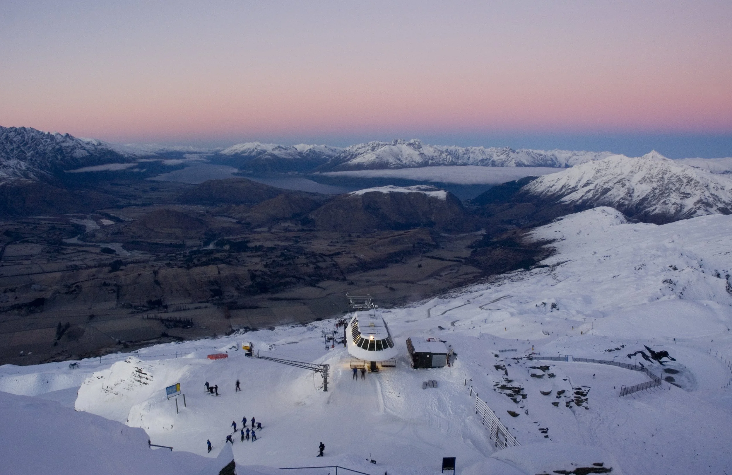 Snow-covered ski station on a mountain with people and a chairlift, overlooking a valley and snow-capped mountains at sunset.