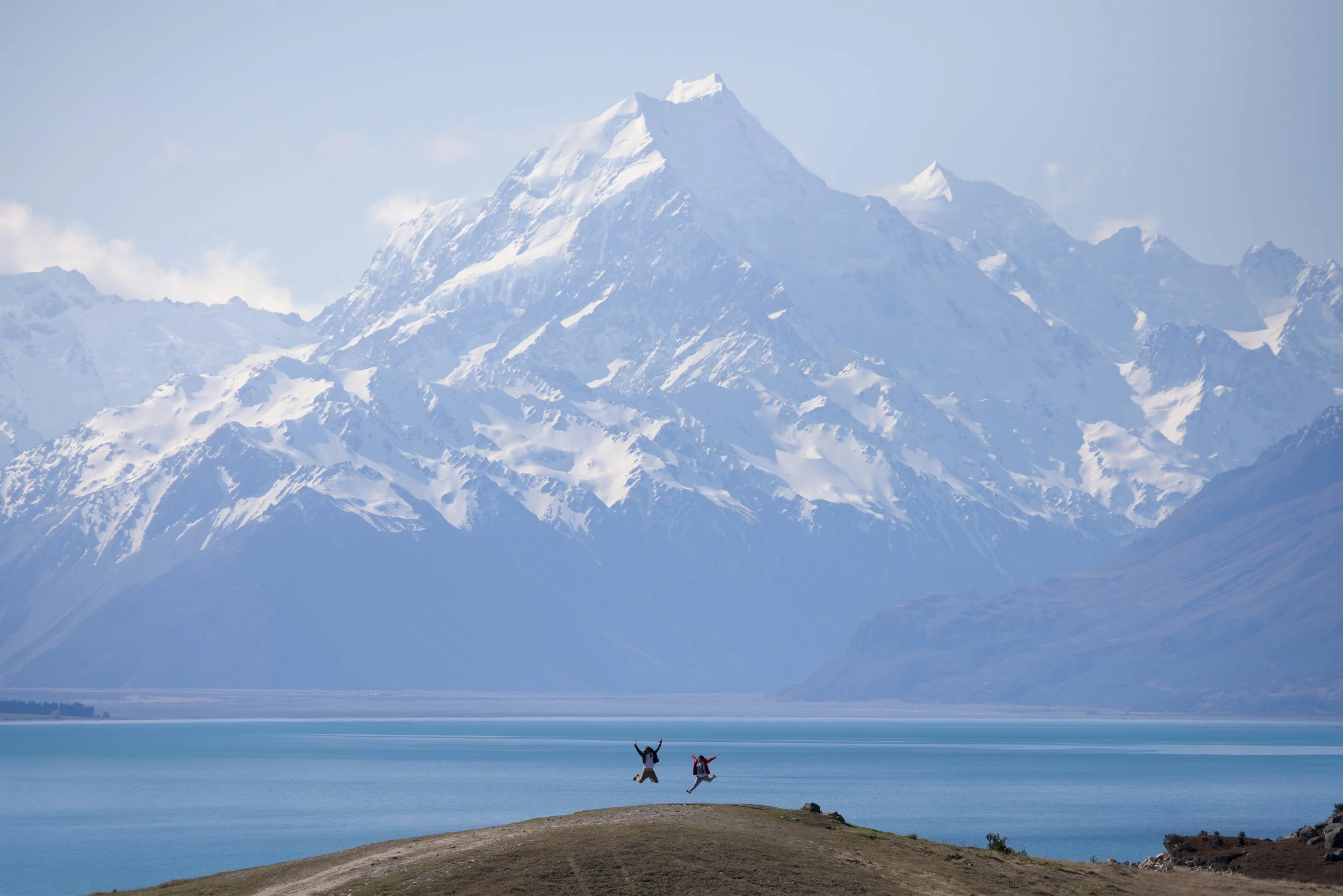 Wide view of two people jumping in front of Lake Pukaki