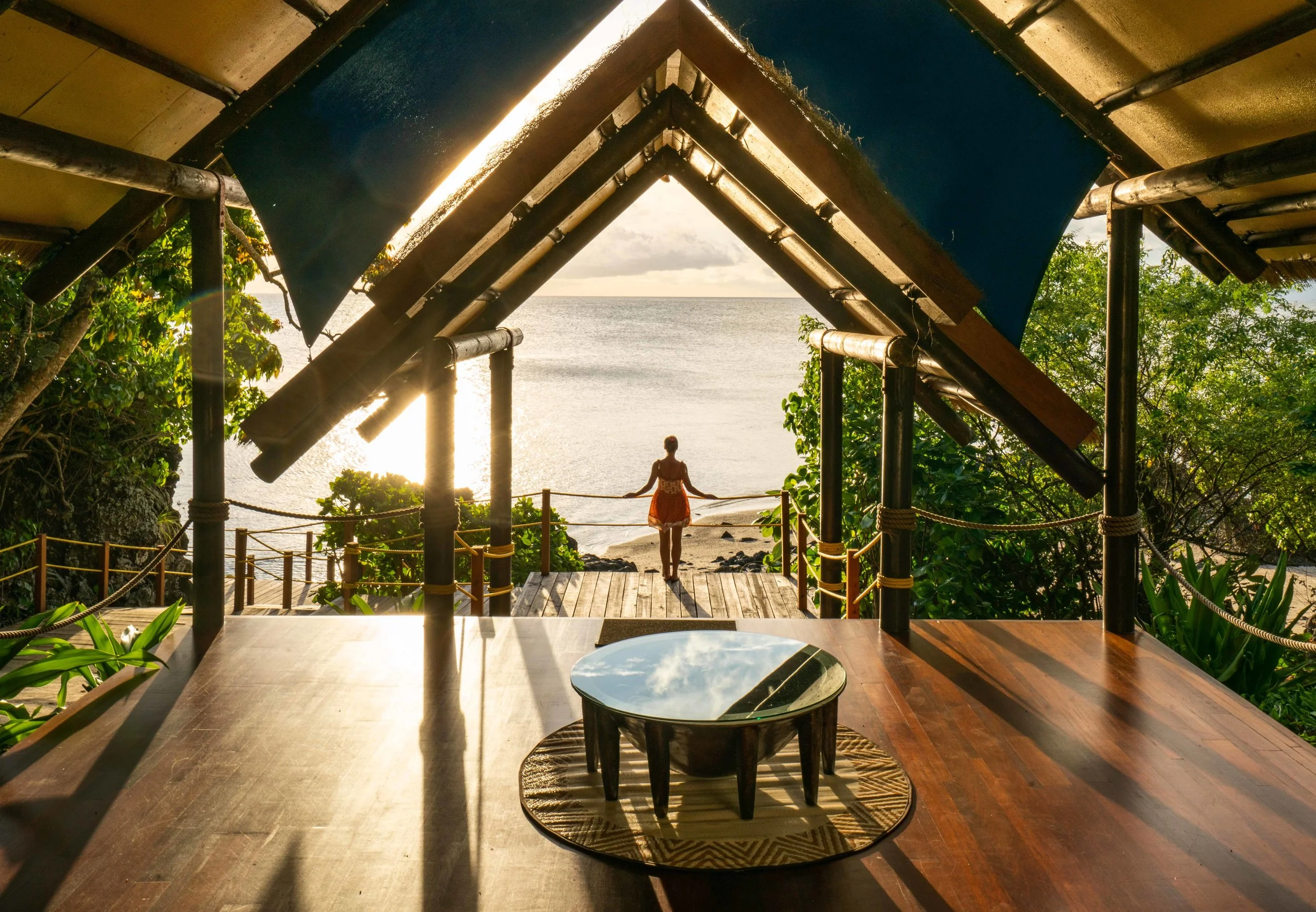 View from inside a thatched-roof hut looking out to a woman standing on a wooden deck near the beach, with the ocean and a partly cloudy sky in the background. The hut has a round table with a reflective surface, and lush green foliage surrounds the scene.