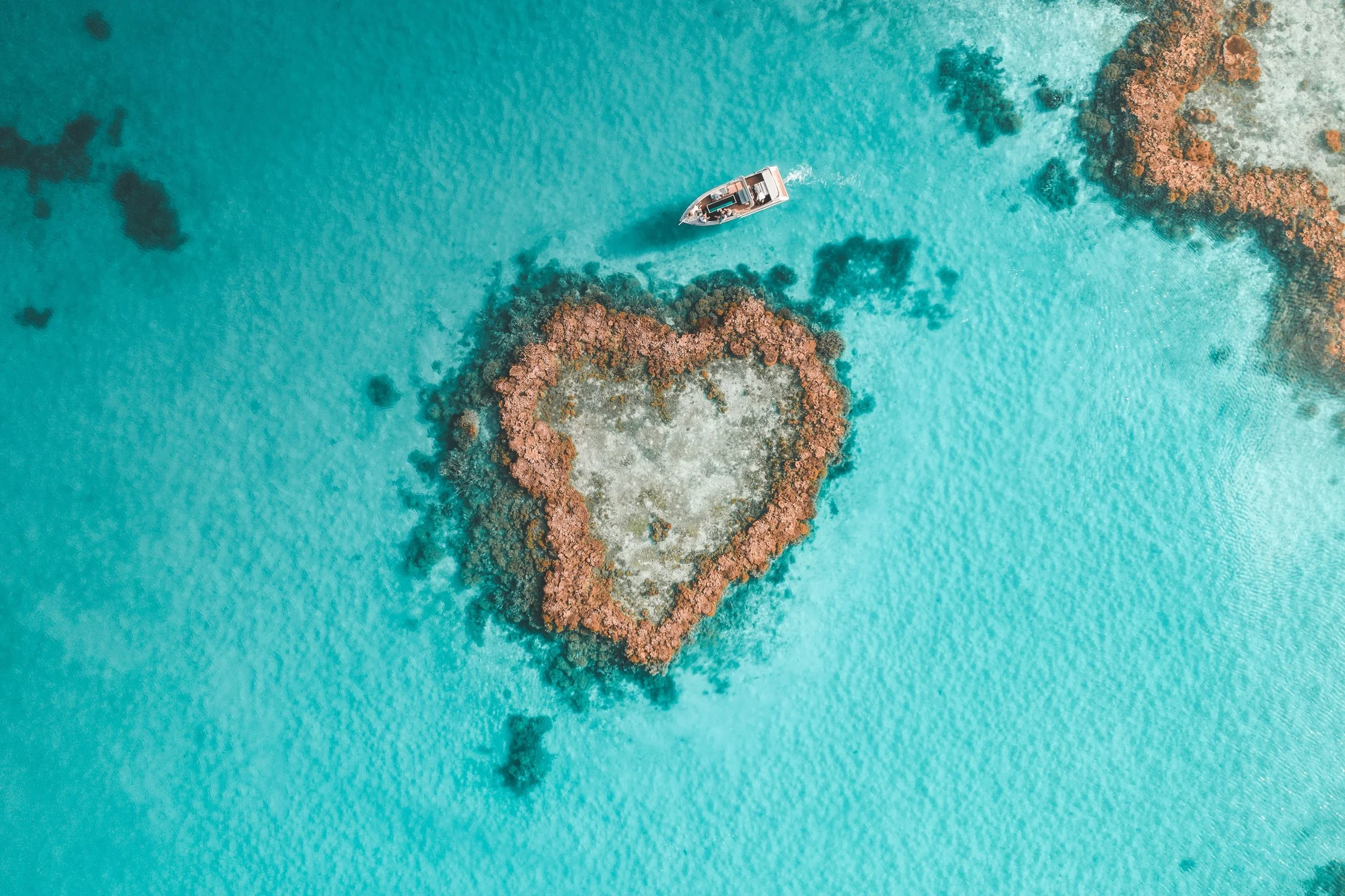 An aerial view of a small boat floating near a heart-shaped coral reef in turquoise water.