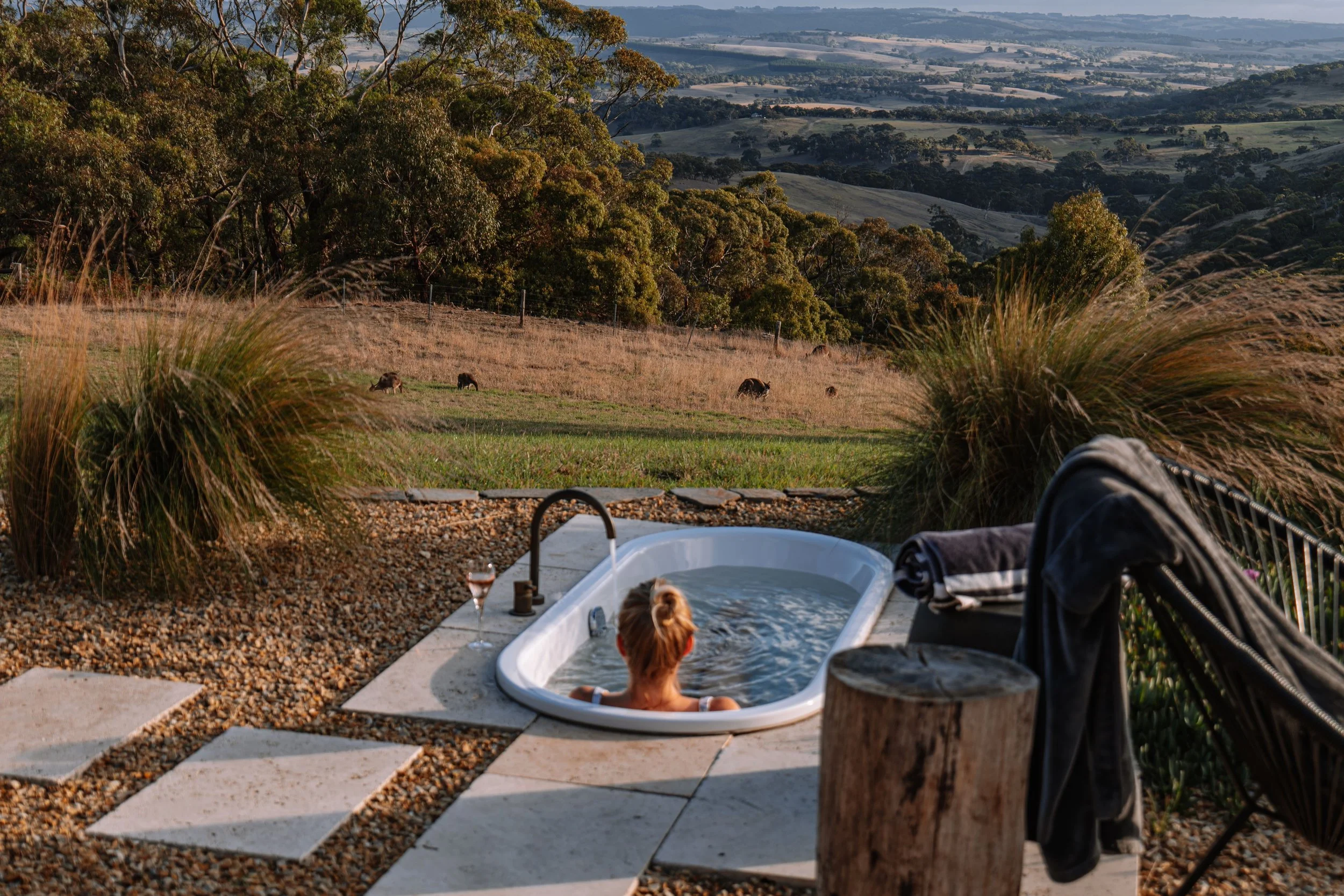 A woman relaxing in an outdoor hot tub with a scenic countryside view of rolling hills and grazing animals in the background, with a glass of wine on the edge of the tub.