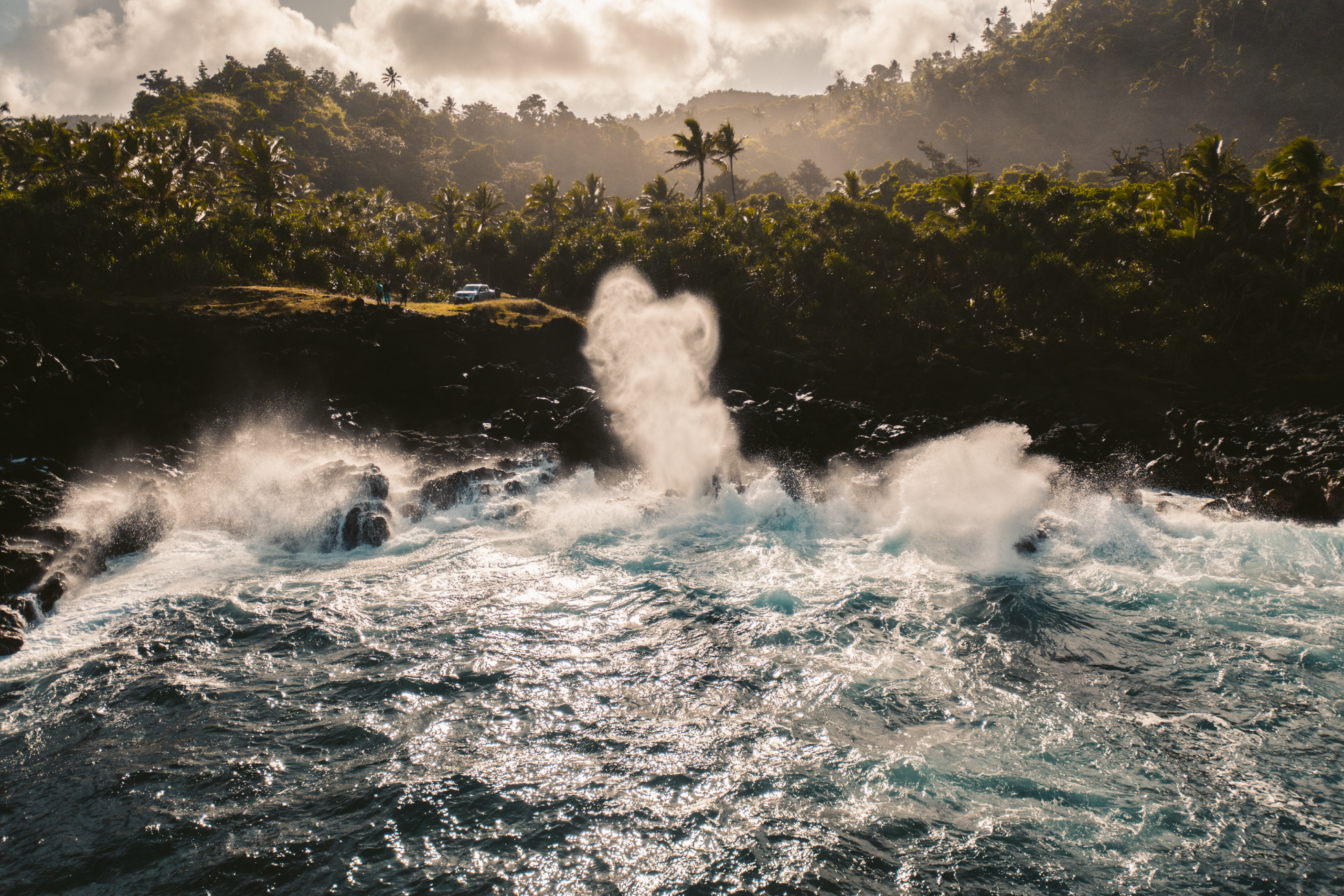Ocean waves crashing onto rocky shore with spray in the air, lush green forested hills in the background under cloudy sky.