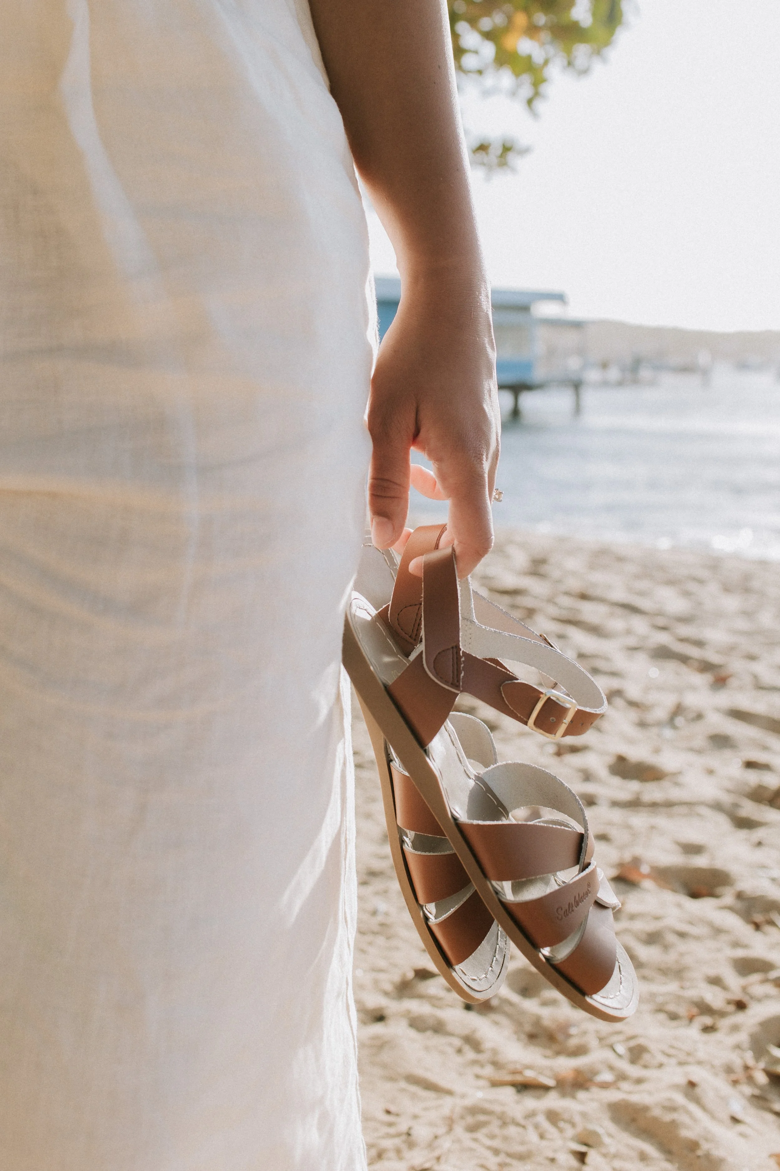 A person holding tan and white sandals on a sandy beach with a pier and water in the background.