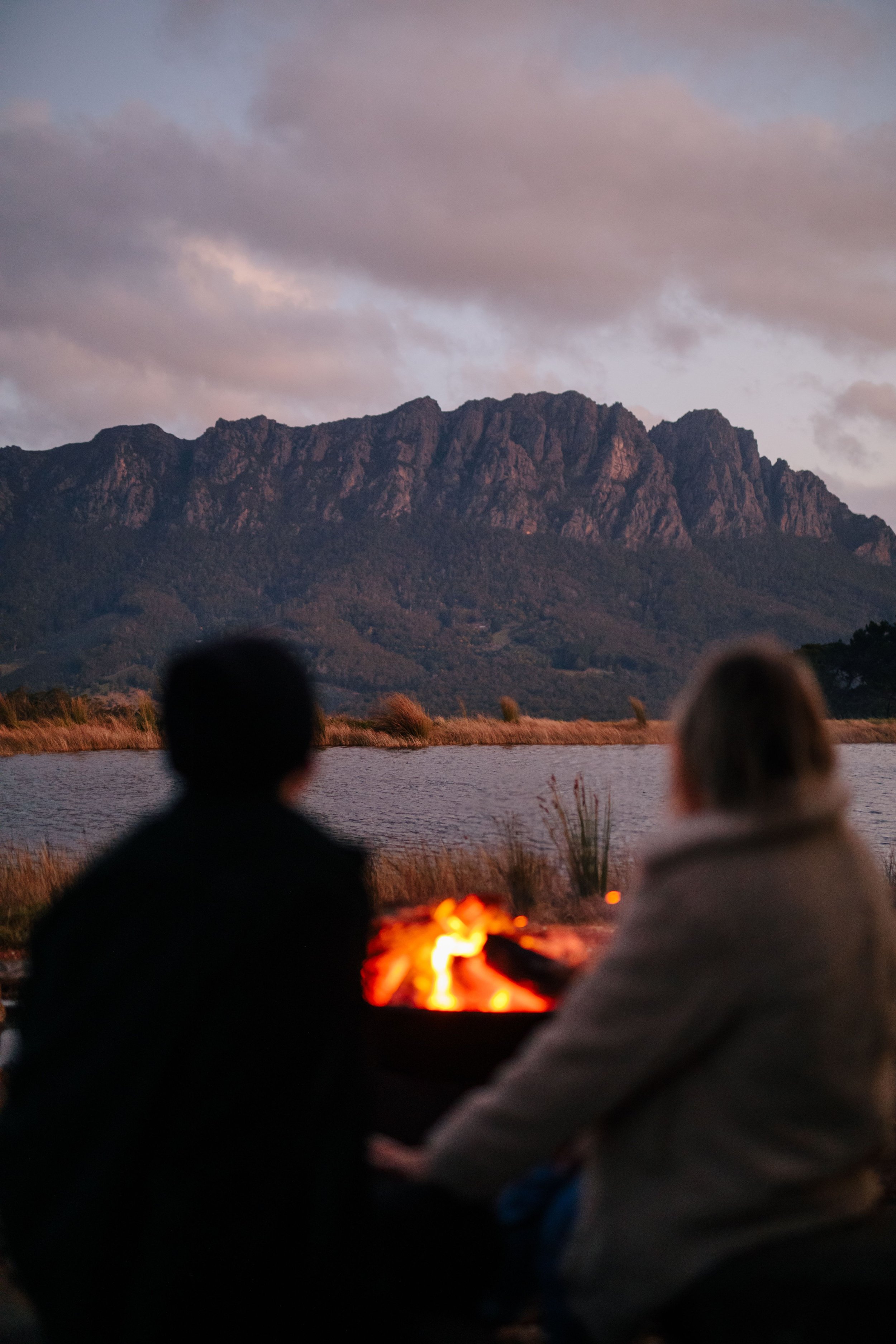 Couple in front of outdoor fire looking out to lake and mountain backdrop