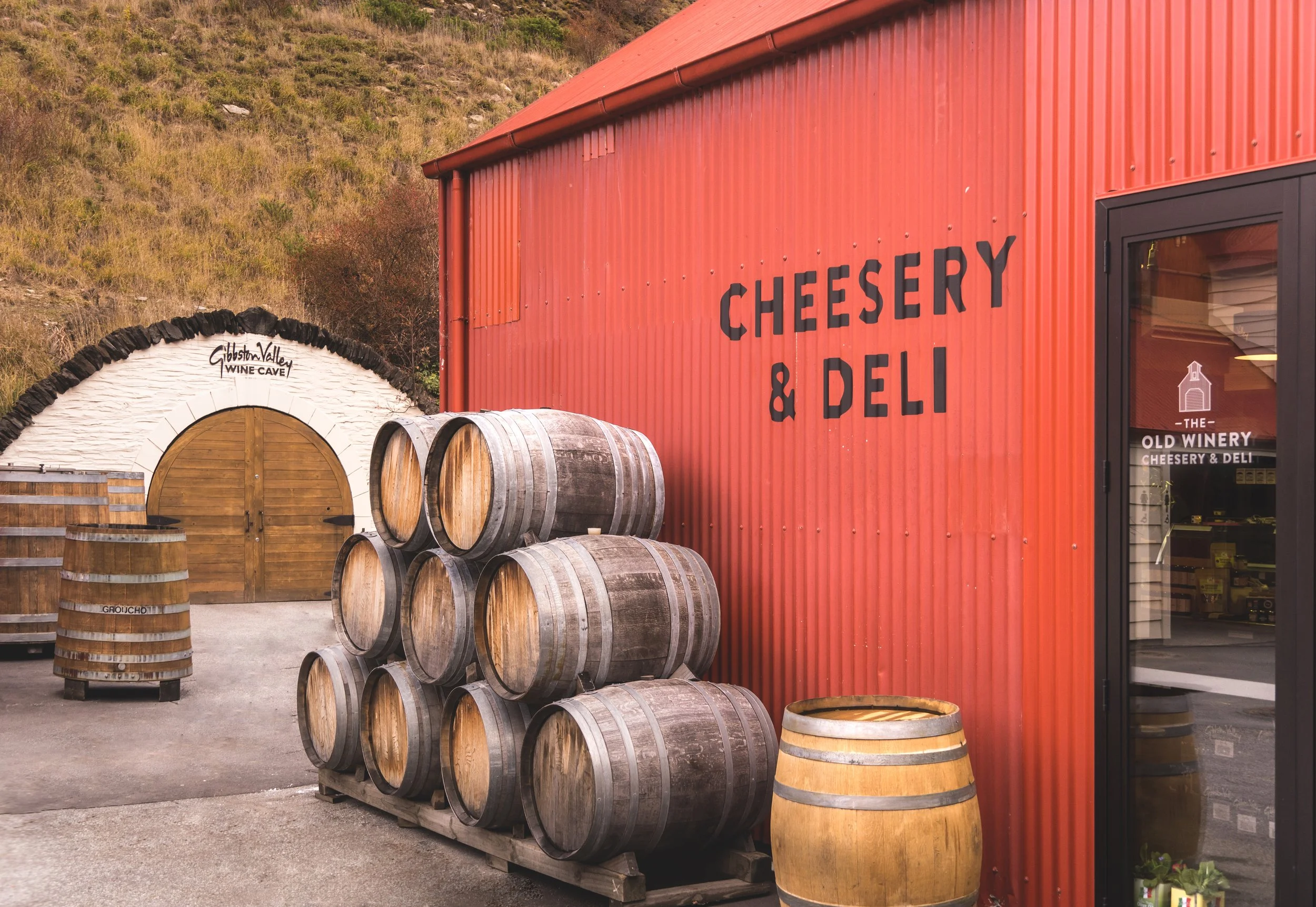 The exterior of a Cheesery & Deli with stacked wine barrels in front, a red corrugated metal building, and a wine cave in the background, located at Gibsonton Valley.