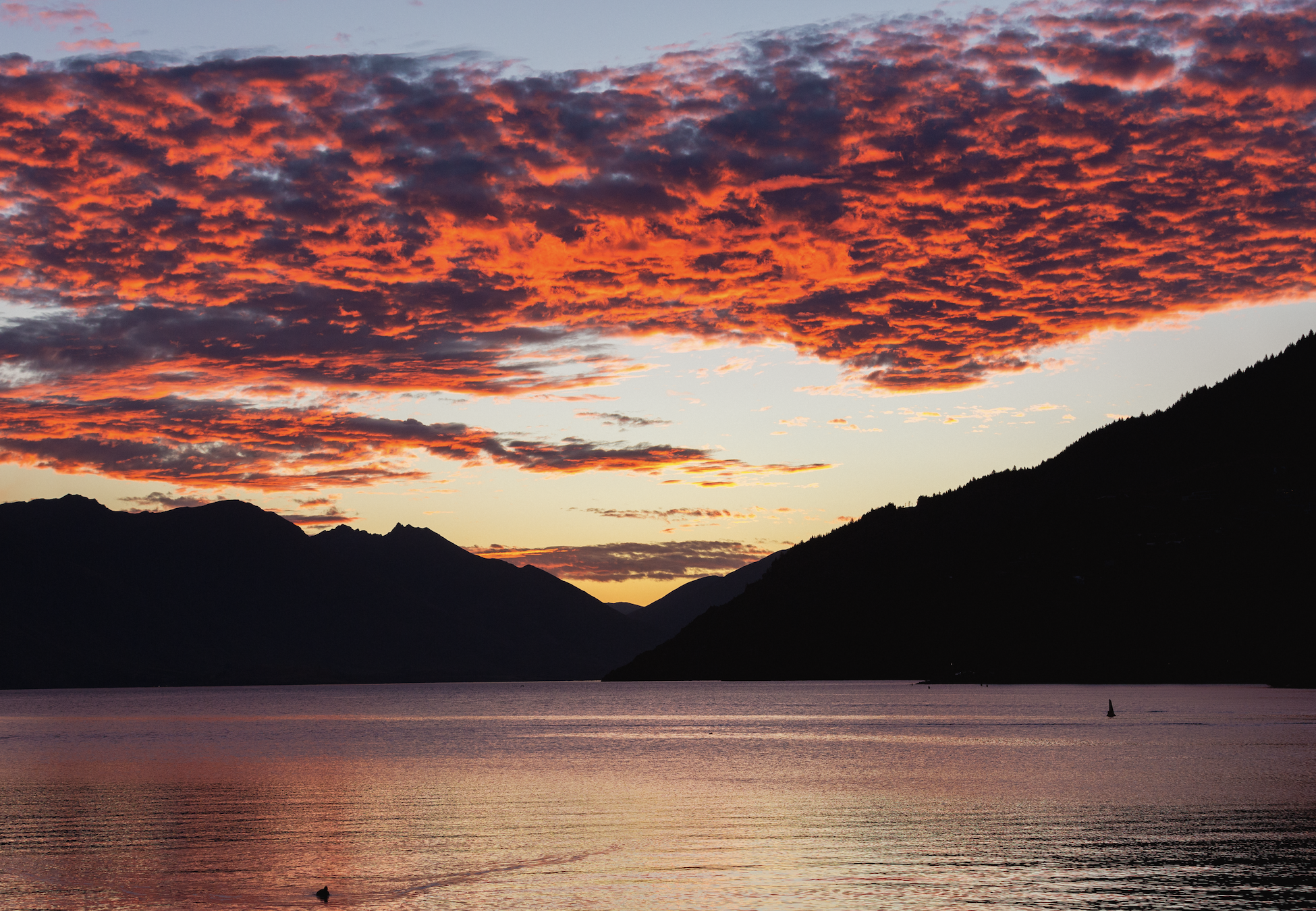 Still lake framed by dark mountain silhouettes under dramatic red sunset clouds