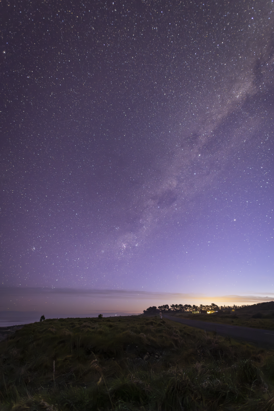 Night sky filled with stars and the Milky Way galaxy over a coastal landscape with trees, a road, and distant lights.