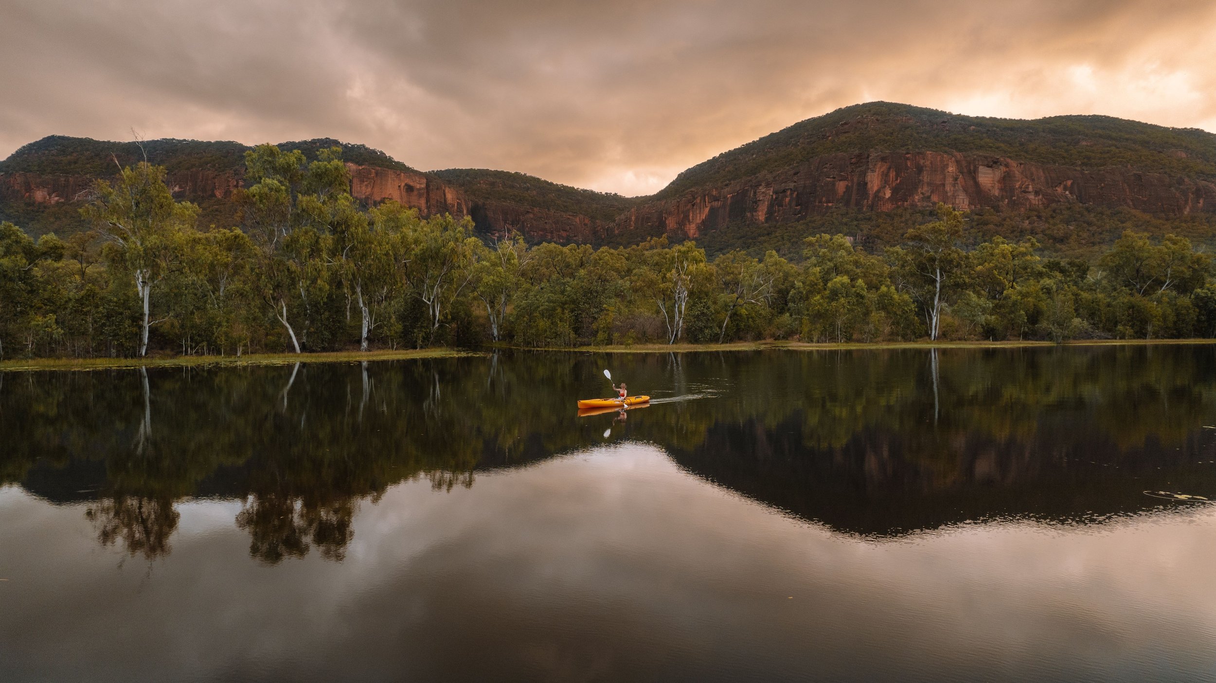 Person kayaking on calm water, surrounded by green trees and mountains under a cloudy sky.