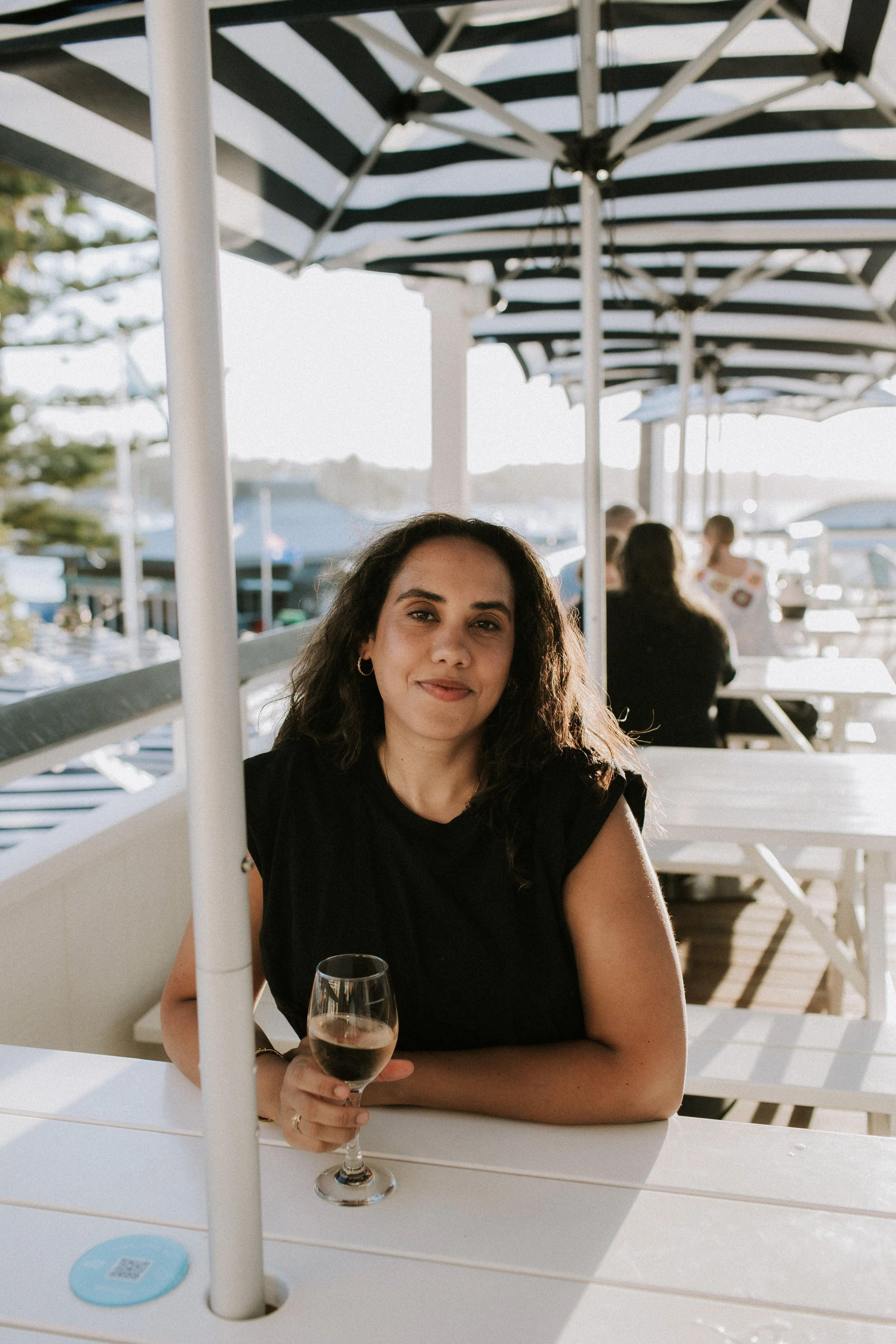Woman sitting at a white table outdoors, holding a glass of rosé wine, at a seaside restaurant with blue and white striped umbrellas and a marina in the background.