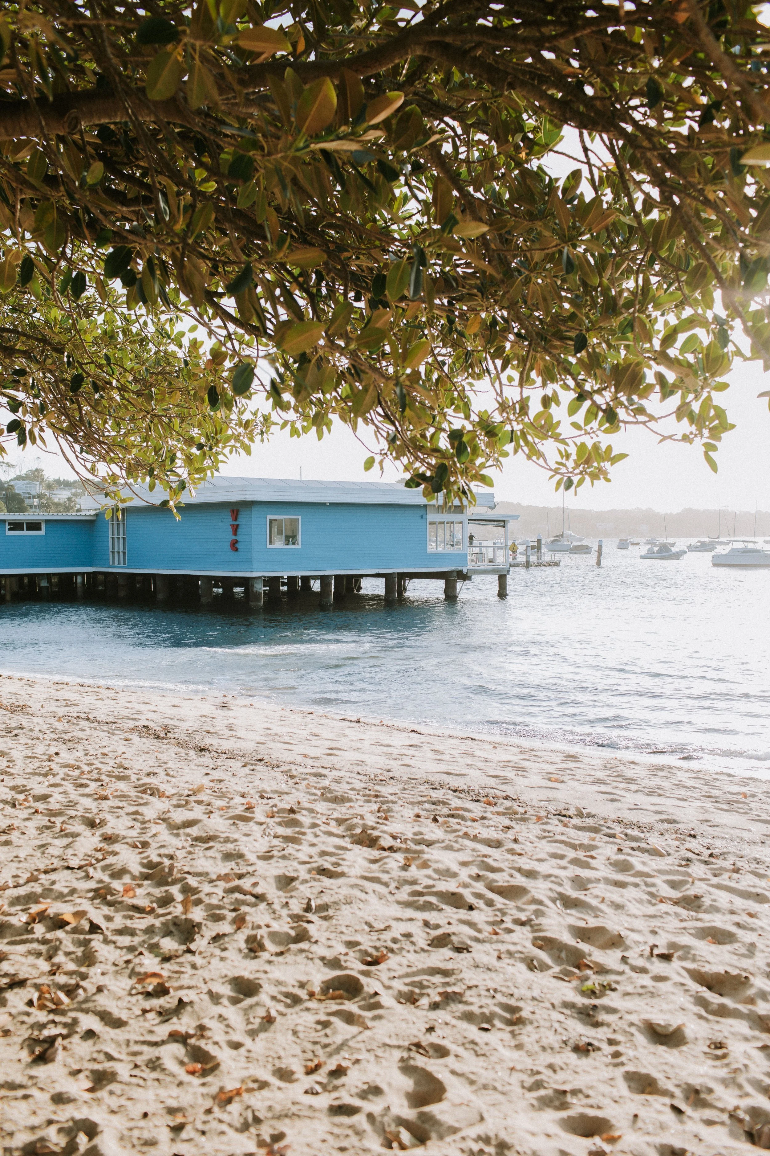 A sandy beach with footprints, looking out to a calm body of water with boats docked, a blue building on stilts, and tree branches overhead, in bright daylight.