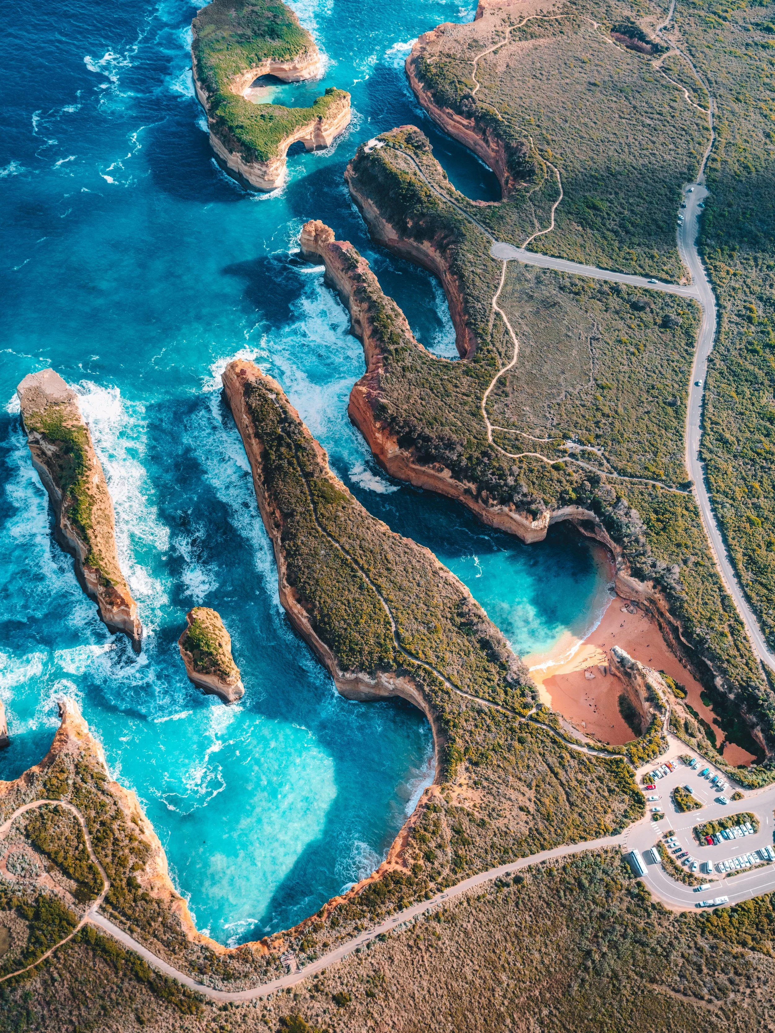 An aerial view of a coastal landscape with turquoise water, rocky cliffs, natural arches, and a parking lot.