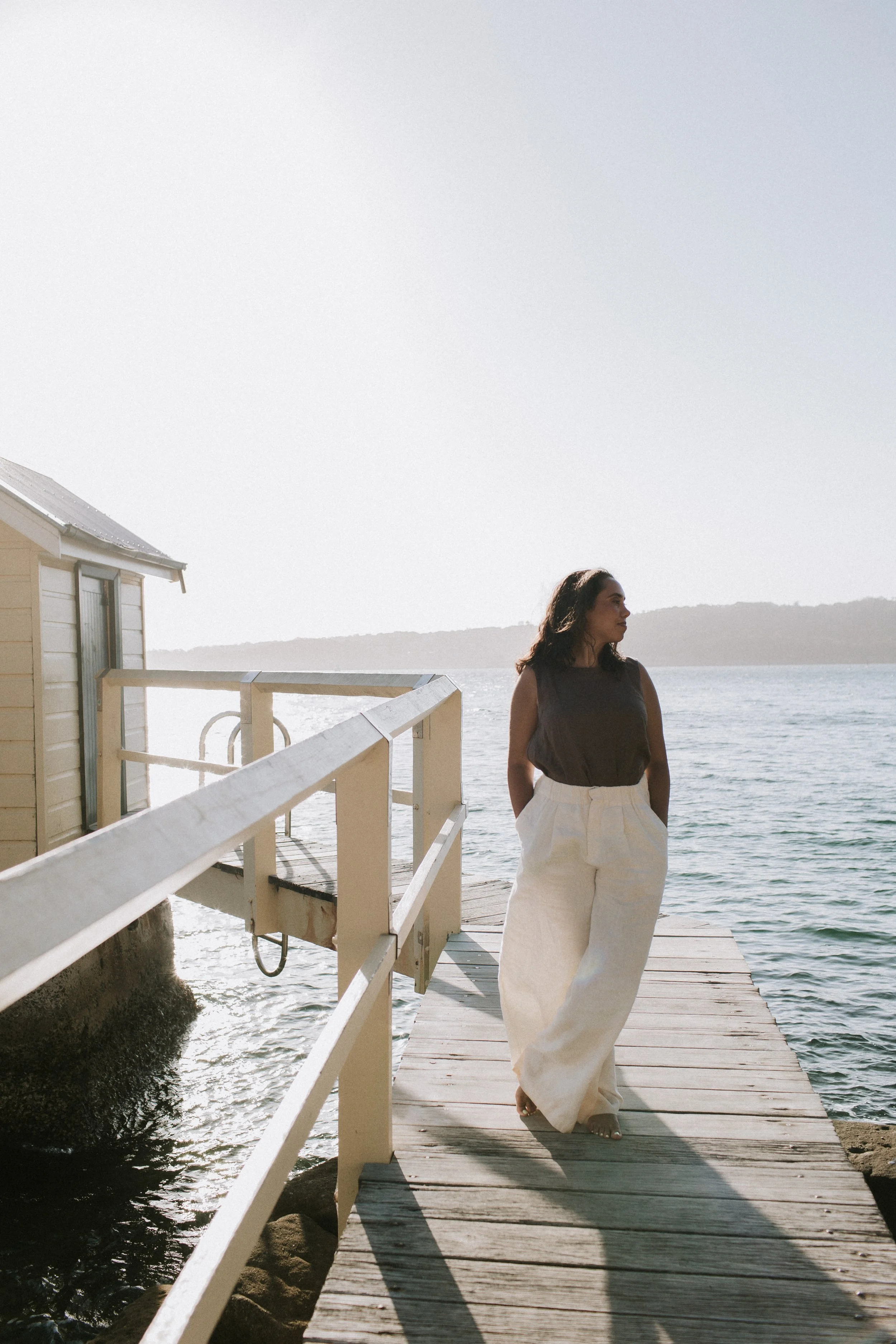 Woman standing on wooden dock by the water, wearing a sleeveless dark top and wide-legged white pants, looking to the side during daytime.