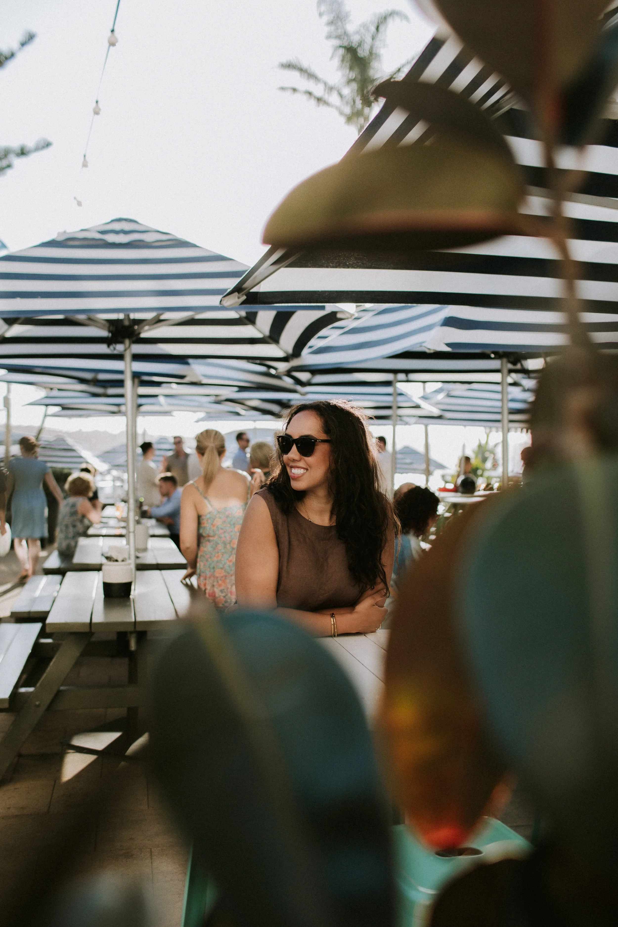 A woman wearing sunglasses and a sleeveless brown top sitting at an outdoor table under blue and white striped umbrellas, smiling and looking to the side with other people in the background.