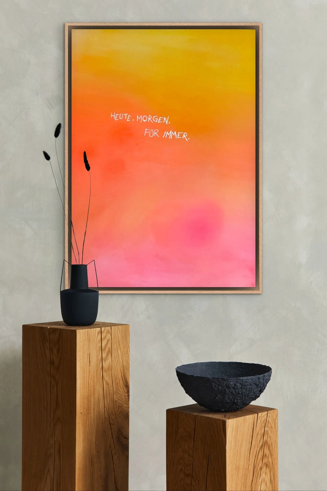 Ein Wandbild mit warmen Farbverläufen in Gelb, Orange und Pink, mit dem weißen Text 'Heute, Morgen, für immer.' in der Mitte. Darunter steht eine schwarze Vase mit getrockneten Gräsern auf einem hohen Holzsockel, neben einem kleineren Holzschädel mit