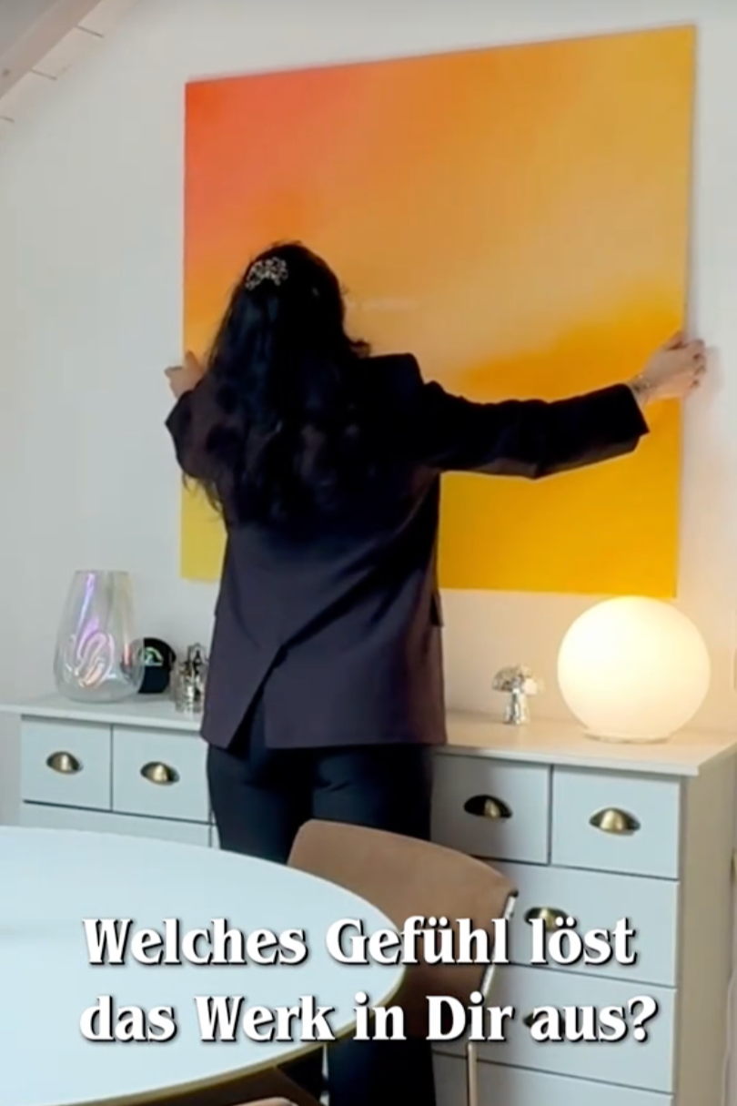 Eine Frau hängt ein großes, teils oranges und gelbes Wandbild auf eine weiße Wand in einem Raum mit weißen Möbeln, einem runden Lampenschirm und Dekorationsgegenständen auf einer Kommode.