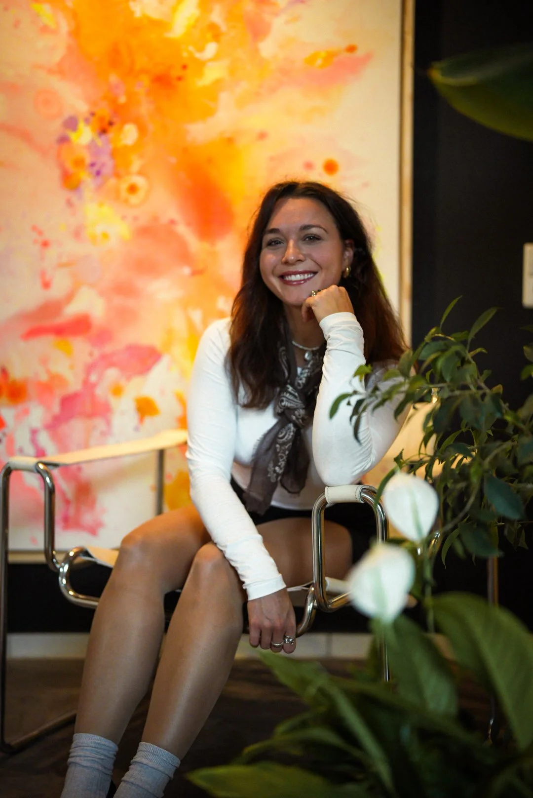 Eine lächelnde Frau sitzt auf einem modernen Sessel, trägt weiße Kleidung, einen Schal, und hat braune, lockige Haare, umgeben von einem grünen Pflanze, mit einer bunten, abstrakten Kunstdruck im Hintergrund.