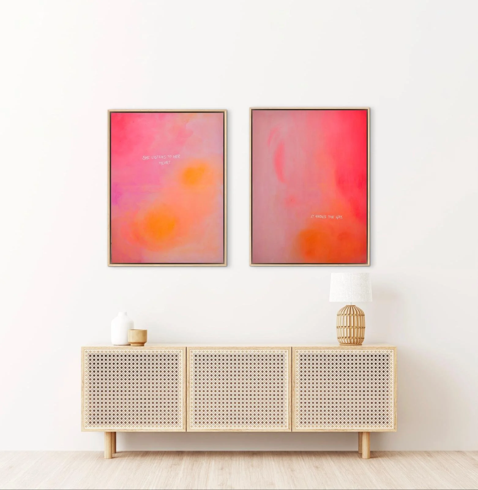Zwei abstrakte Gemälde mit warmen Farben in Rahmen hängen an einer weißen Wand, darunter steht eine helle Kommode mit drei Fächern, auf der mehrere Vasen und eine Lampe stehen.