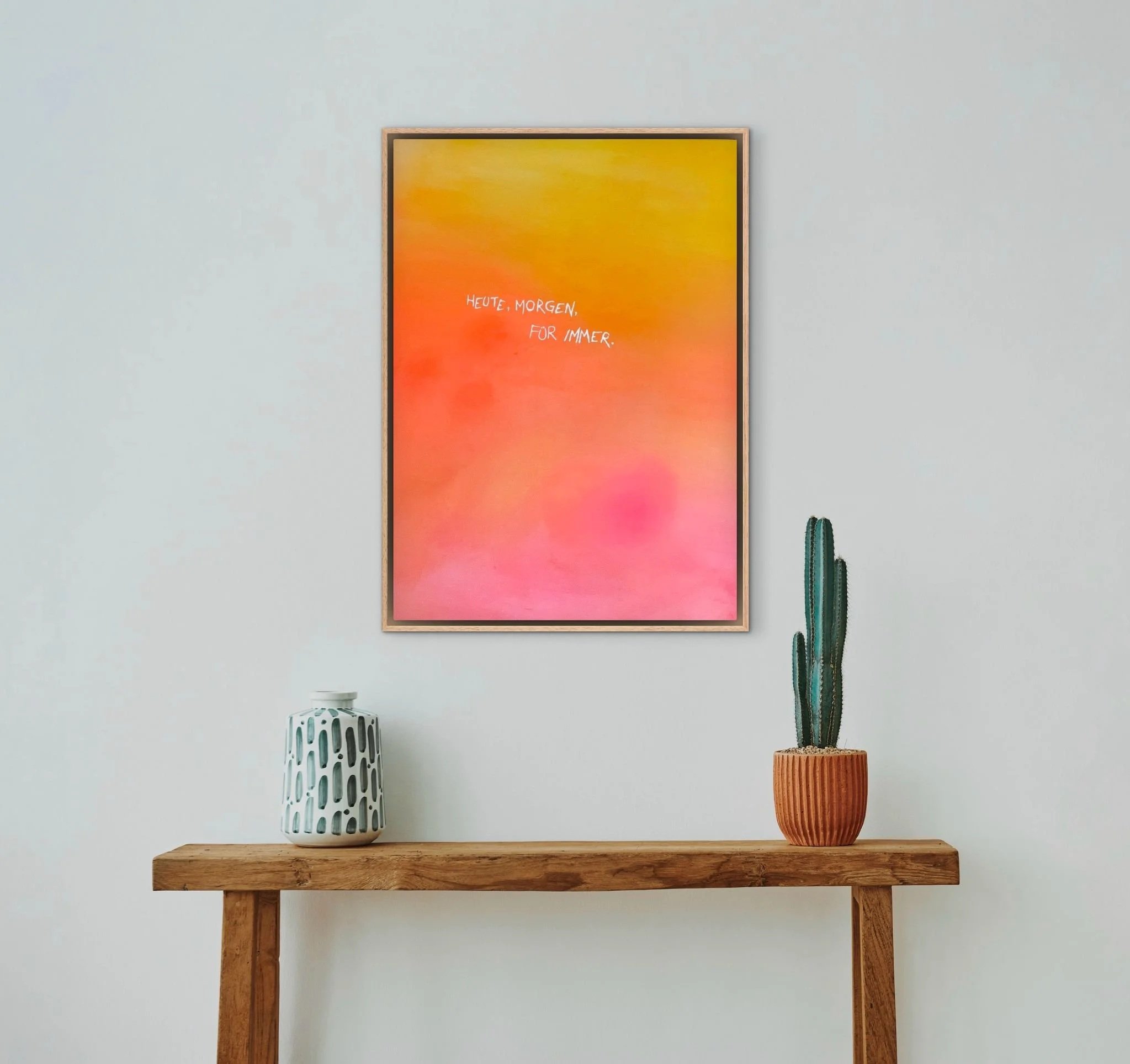 Ein Wandbild mit warmen Farbverlauf von Gelb bis Pink, mit dem Text 'Heute, morgen, für immer'. Darunter ein Holzregal mit einer weißen Vase und einem Topf mit Kaktus.