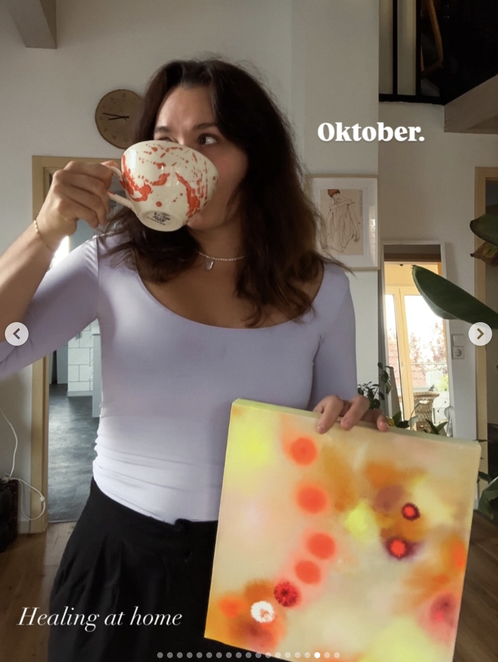 Eine Frau trinkt aus einer weißen Tasse mit rotem Muster, hält ein buntes abstraktes Gemälde in der Hand, im Hintergrund ein Wohnzimmer mit Fenster und Pflanzen, der Text 'Oktober.' und 'Healing at home'.