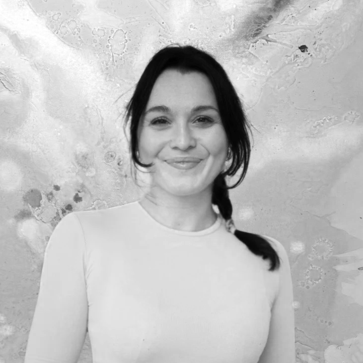 Schwarz-weiß-Foto einer lächelnden jungen Frau mit dunklen Haaren, die eine weiße Oberbekleidung trägt, vor einer texturierten Wand im Hintergrund.