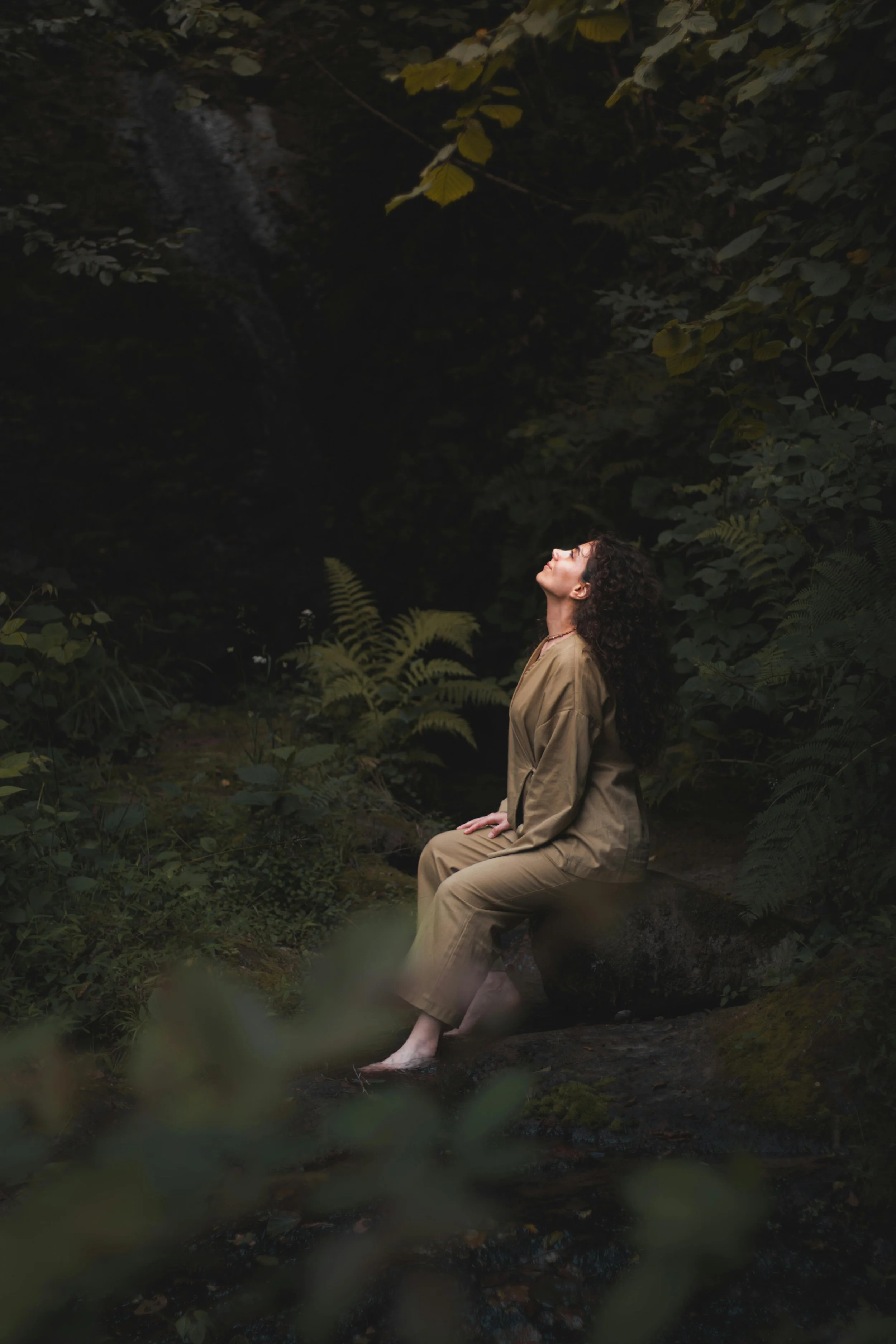 A woman with curly hair sitting on a rock in a lush, green forest, with her feet in the water, and her face turned upward with eyes closed.