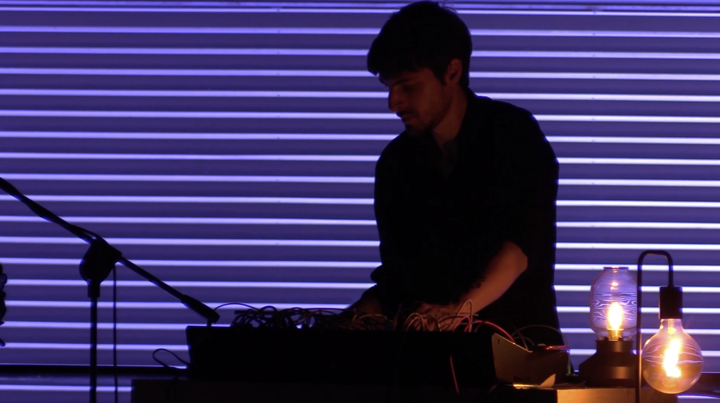 A silhouette of a person operating electronic music equipment against a background of horizontal blue and purple striped lights with a couple of glowing lamps on the right side.