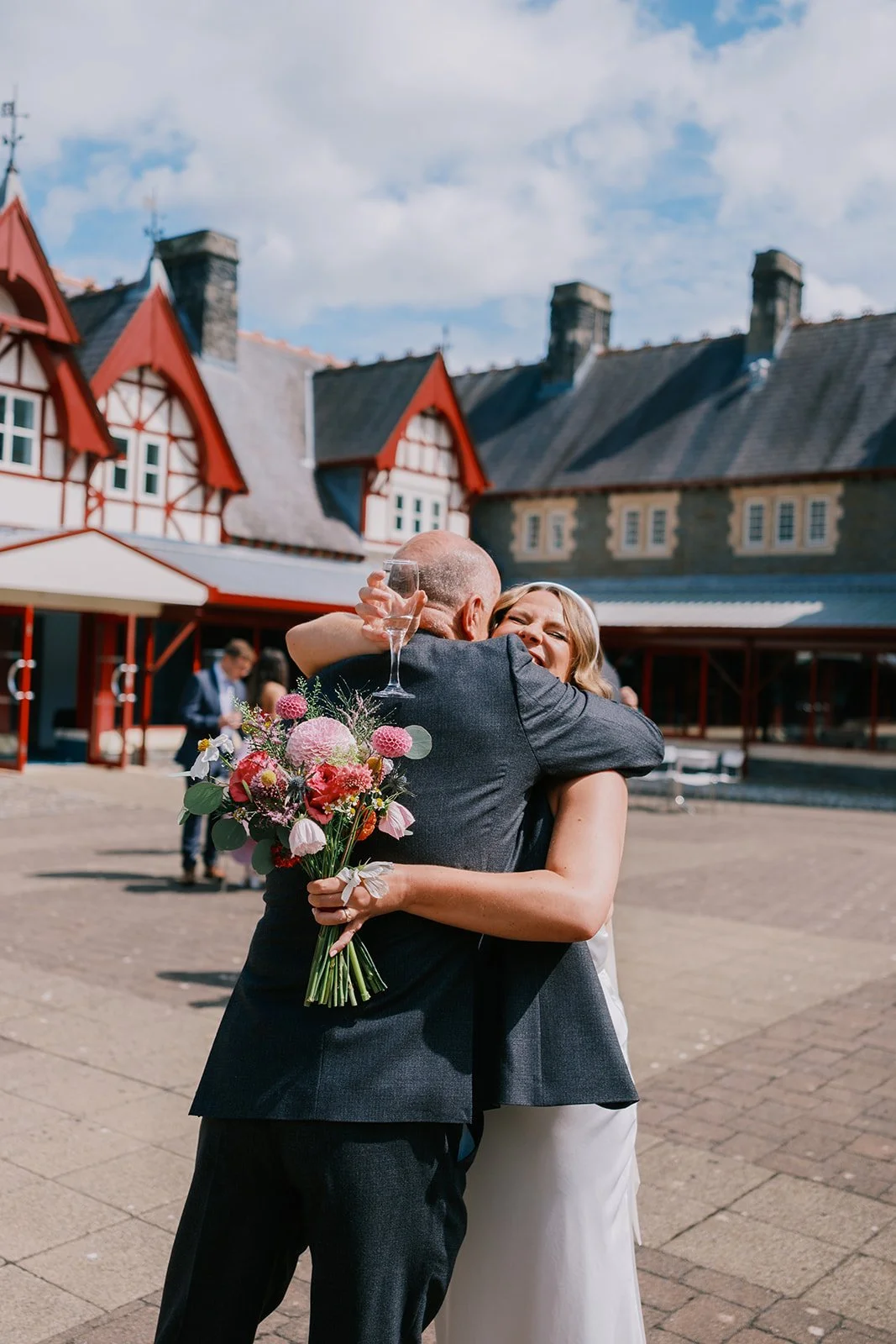 A joyful embrace between a woman in a white dress holding a bouquet of flowers and an older man in a suit holding a champagne glass, set in front of a traditional building with red gabled roofs under a blue sky.