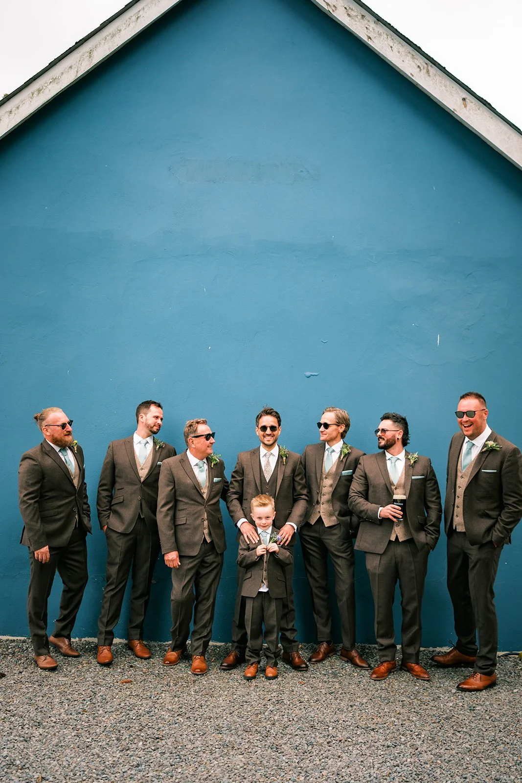 Group of men in suits posing in front of a bright blue wall, with one young boy in the center holding a small bouquet, all smiling and wearing sunglasses.